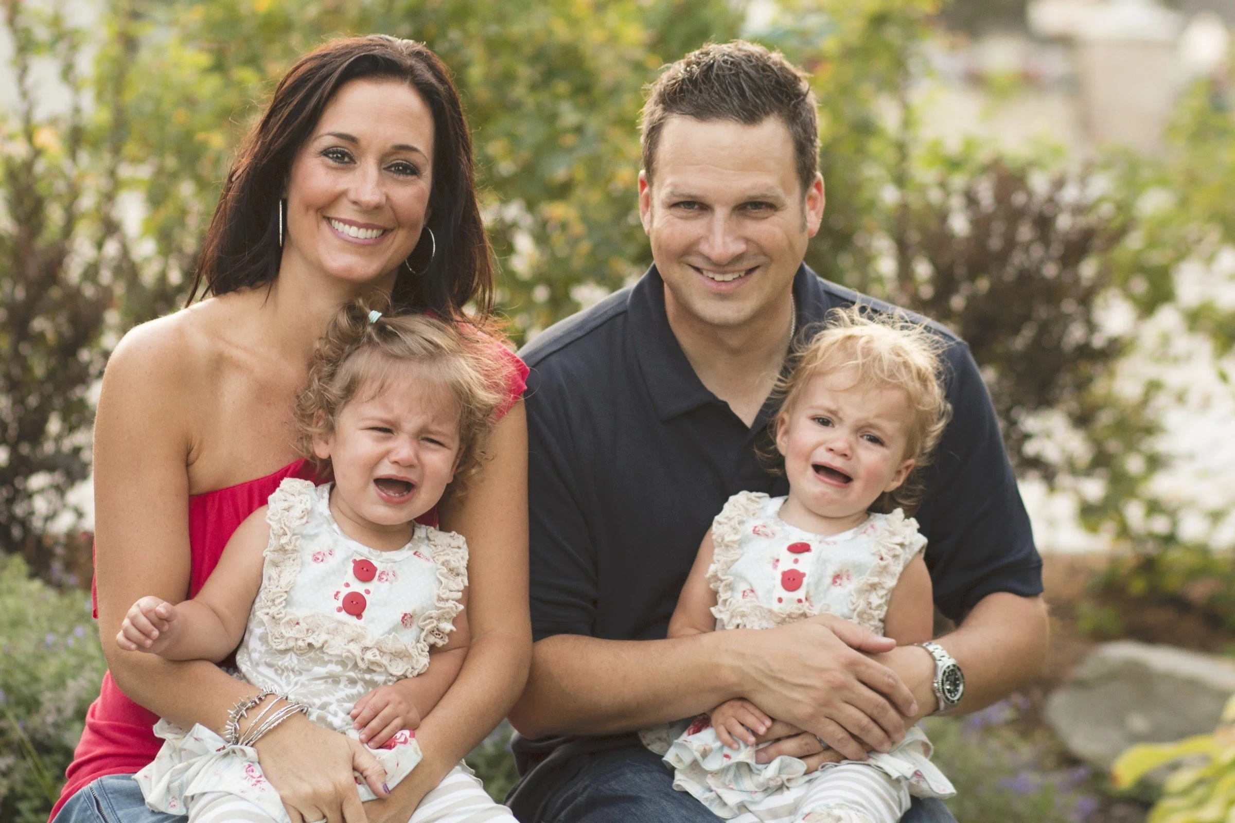 A happy family of four outdoors with two women and two young children, all smiling or crying, in a garden setting with trees and rocks in the background.