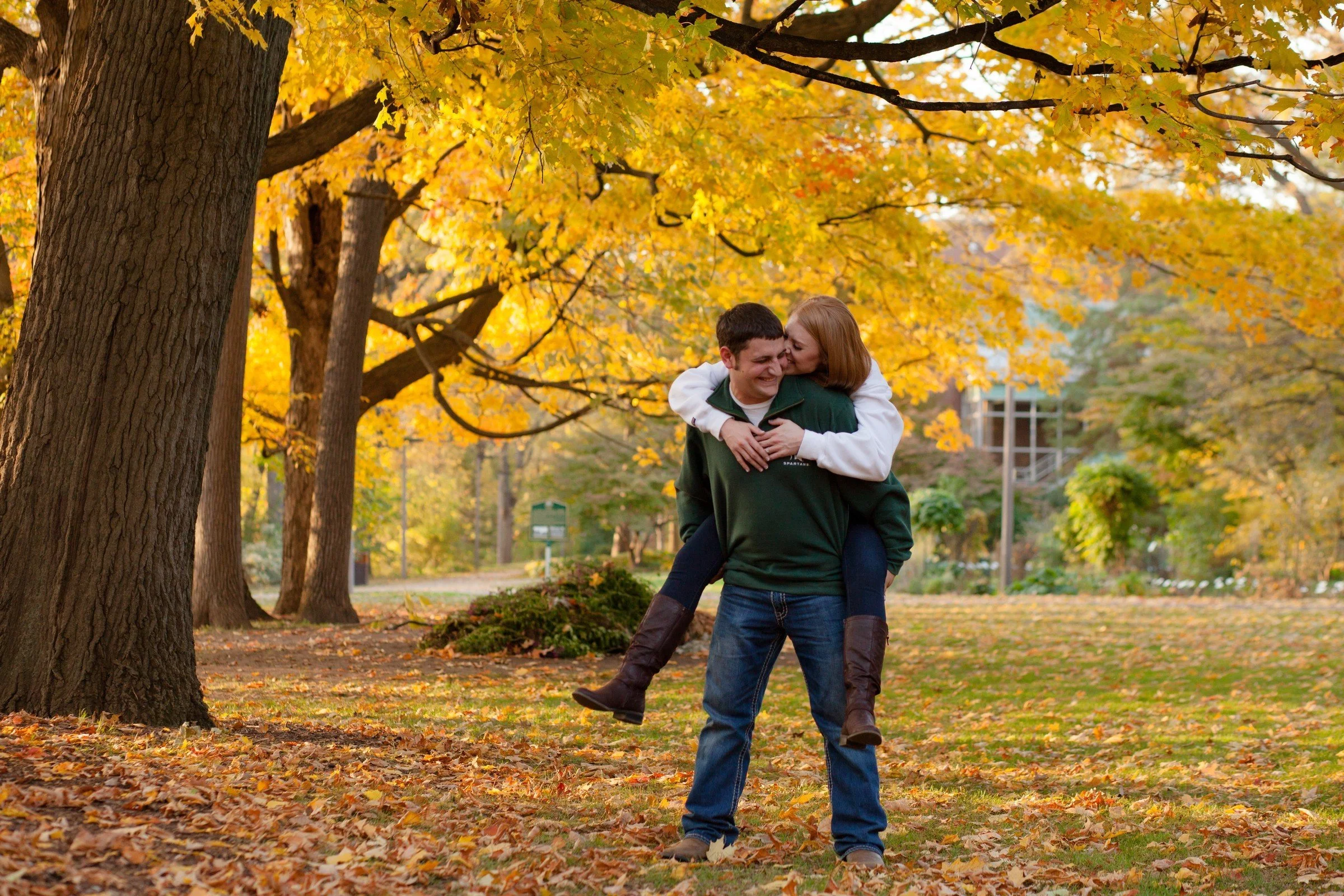 A young couple in a park during autumn, with the woman giving the man a piggyback ride. The trees around them have yellow and orange leaves, and the ground is covered with fallen leaves.