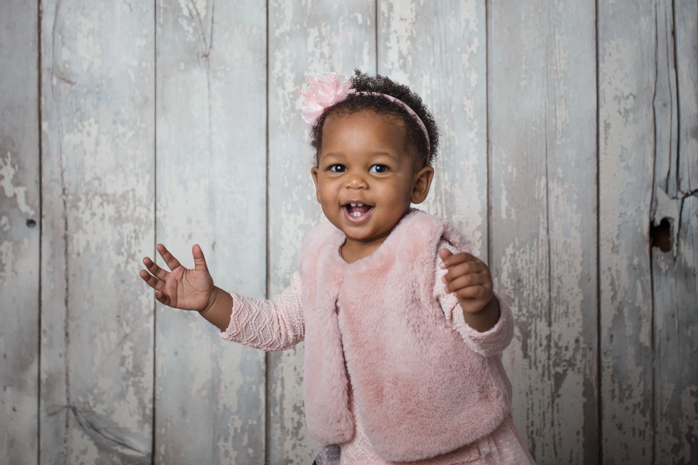 A happy young African American girl with curly hair, wearing a pink headband with a bow, a pink furry vest, and a pink dress, standing in front of a rustic wooden wall.