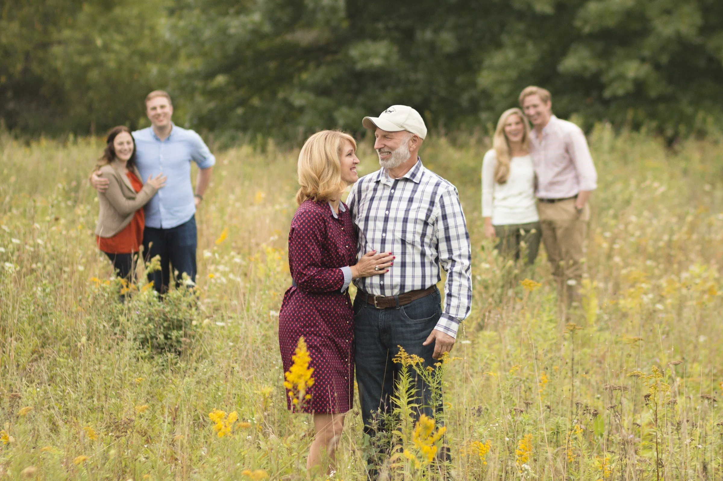 A happy older couple stands close together in a field of wildflowers, smiling and looking into each other's eyes. In the background, two younger couples are also smiling and talking, with trees and greenery behind them.