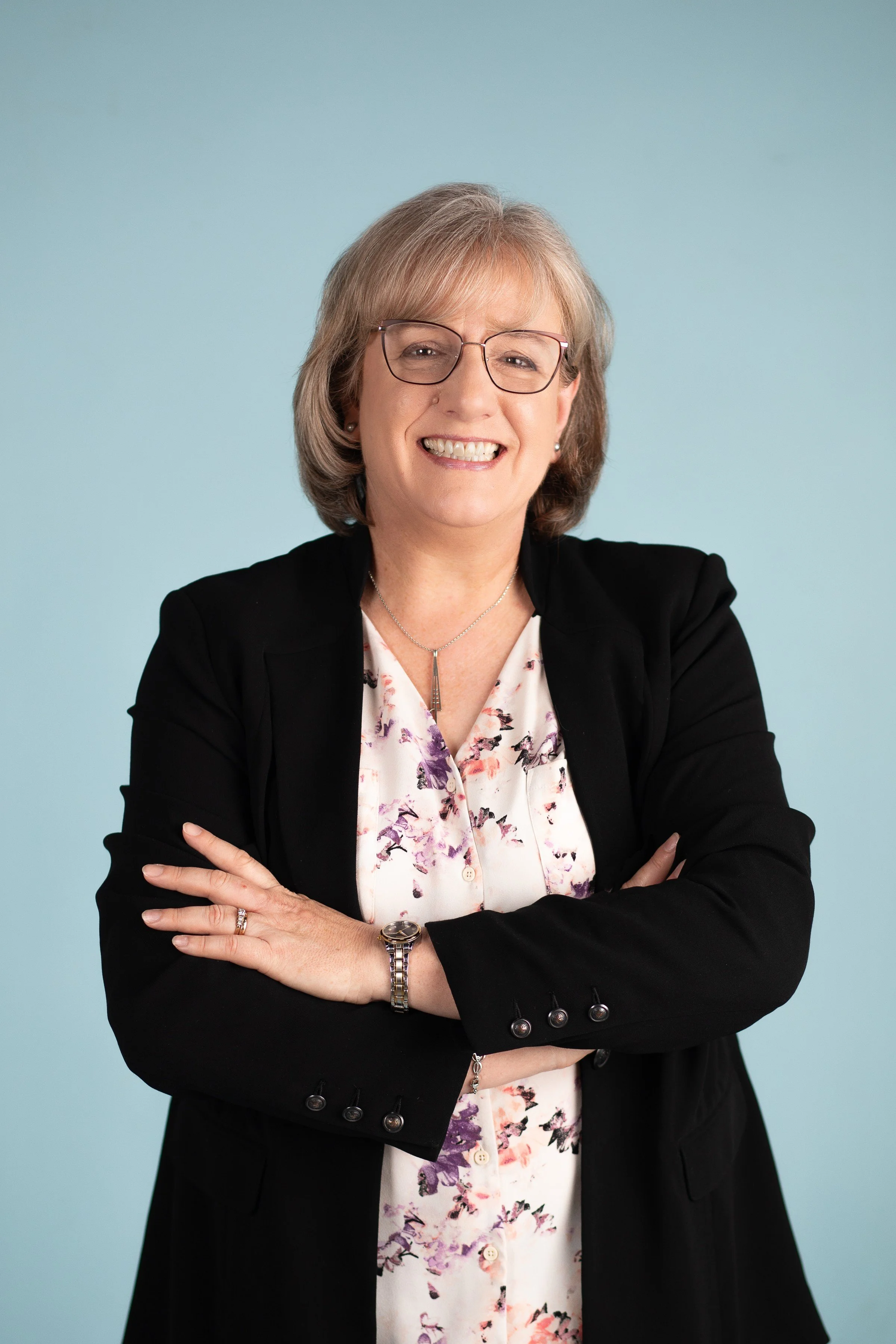 A smiling middle-aged woman with blonde hair, glasses, wearing a black blazer over a floral blouse, standing against a light blue background.