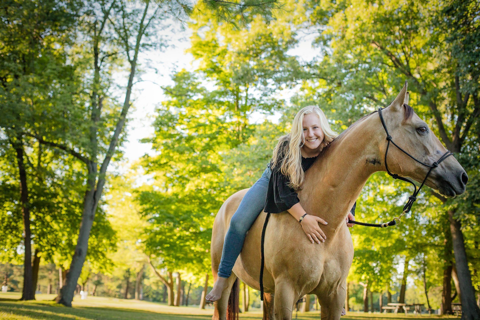 A young woman with blonde hair riding a tan horse in a park with green trees and sunlight.