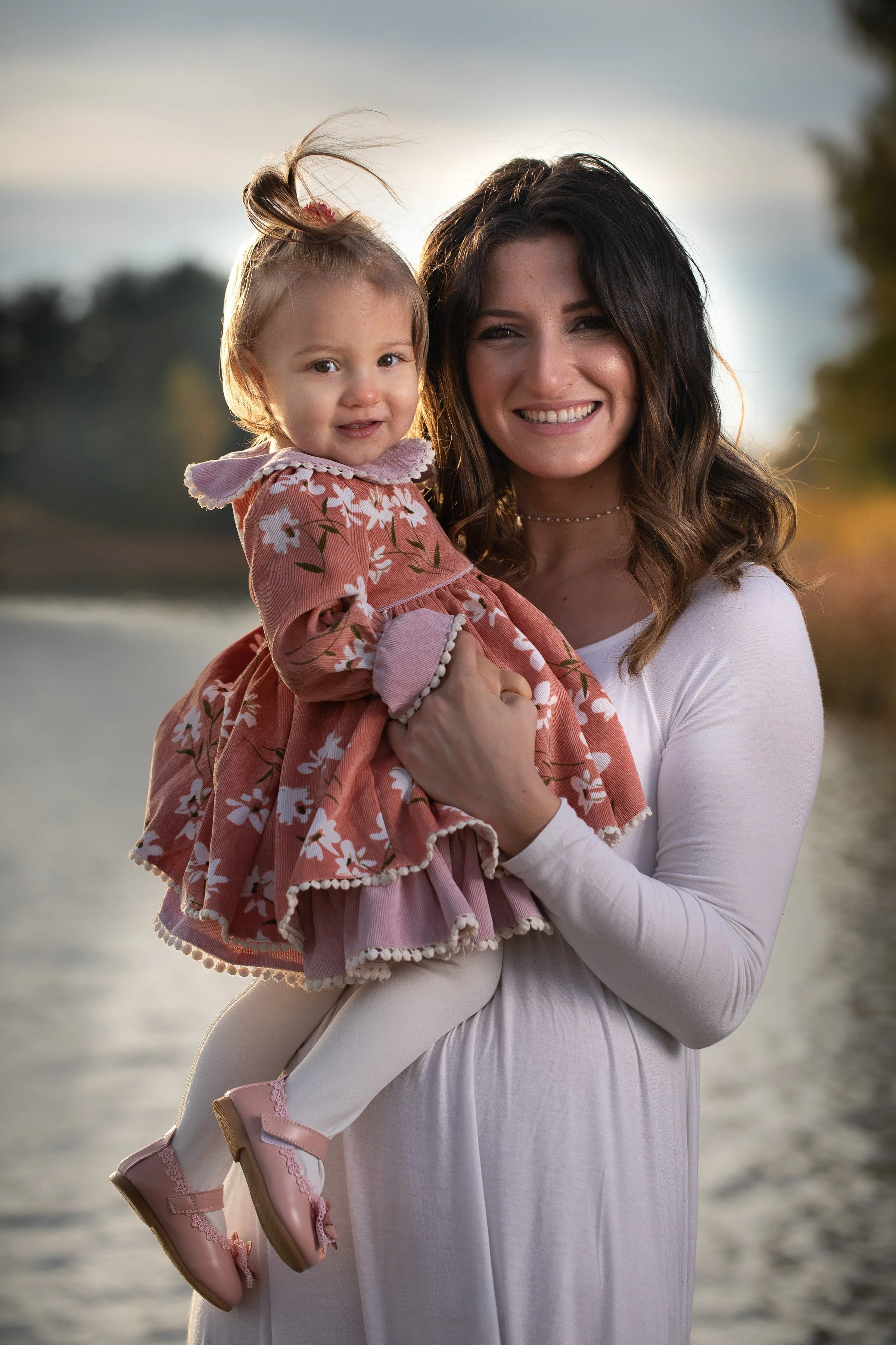 A woman holding a young girl on a lakeside during sunset, both smiling at the camera.