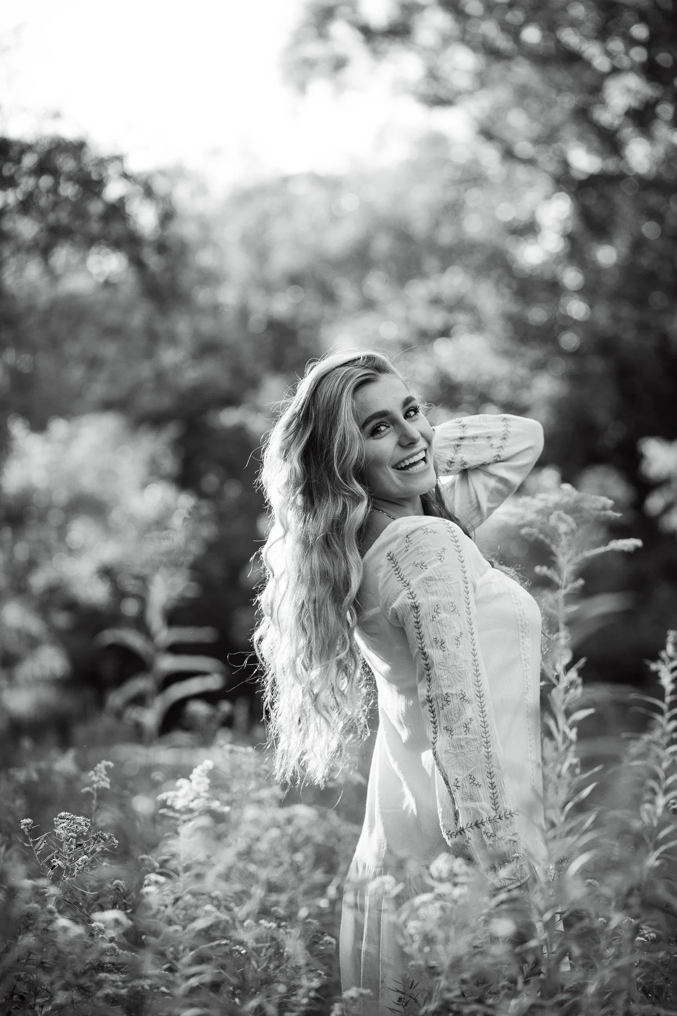 A smiling woman with long, wavy hair standing outdoors in a natural setting with trees and plants, in black and white.
