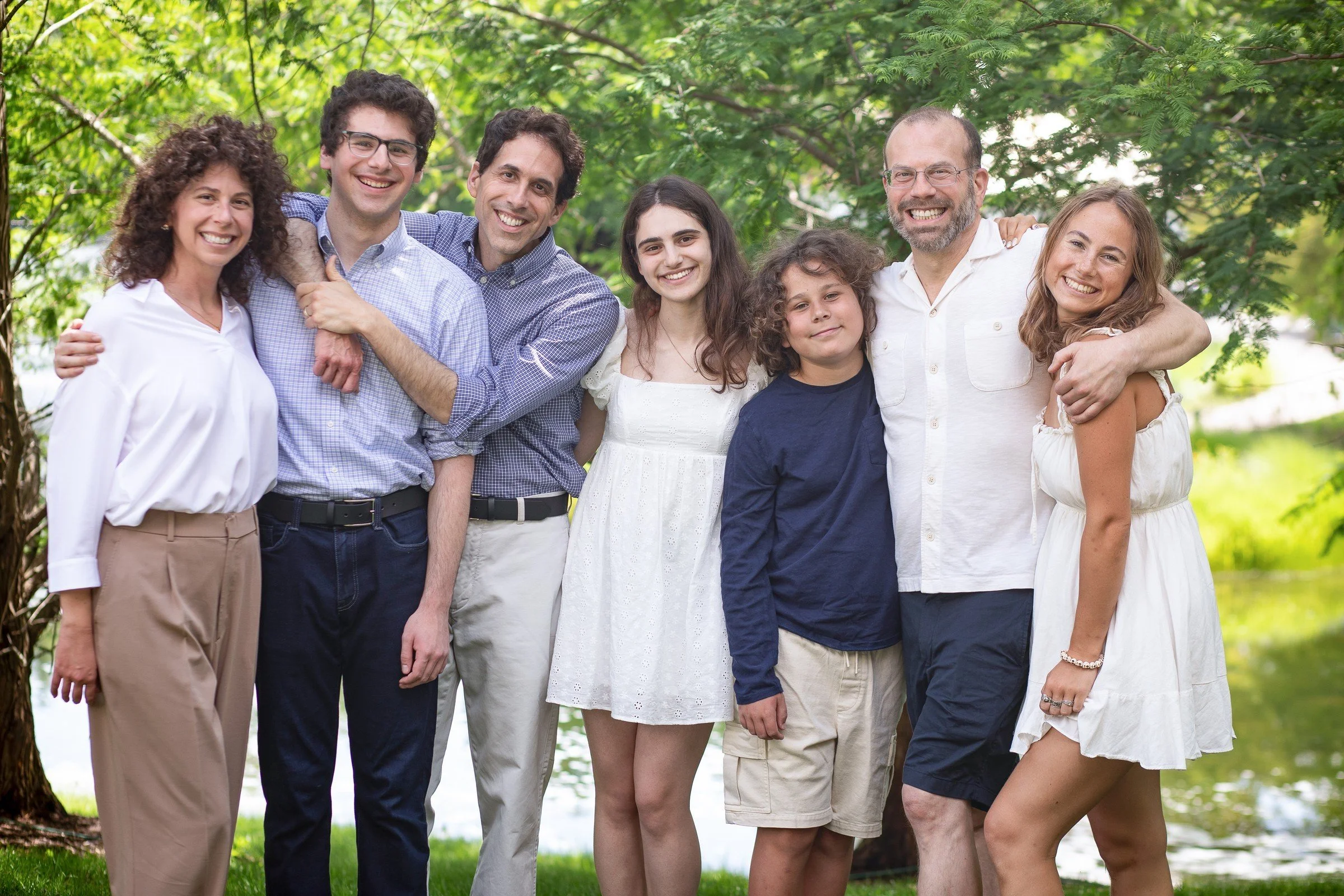 Group of eight people smiling outdoors near a tree and water, embracing each other.