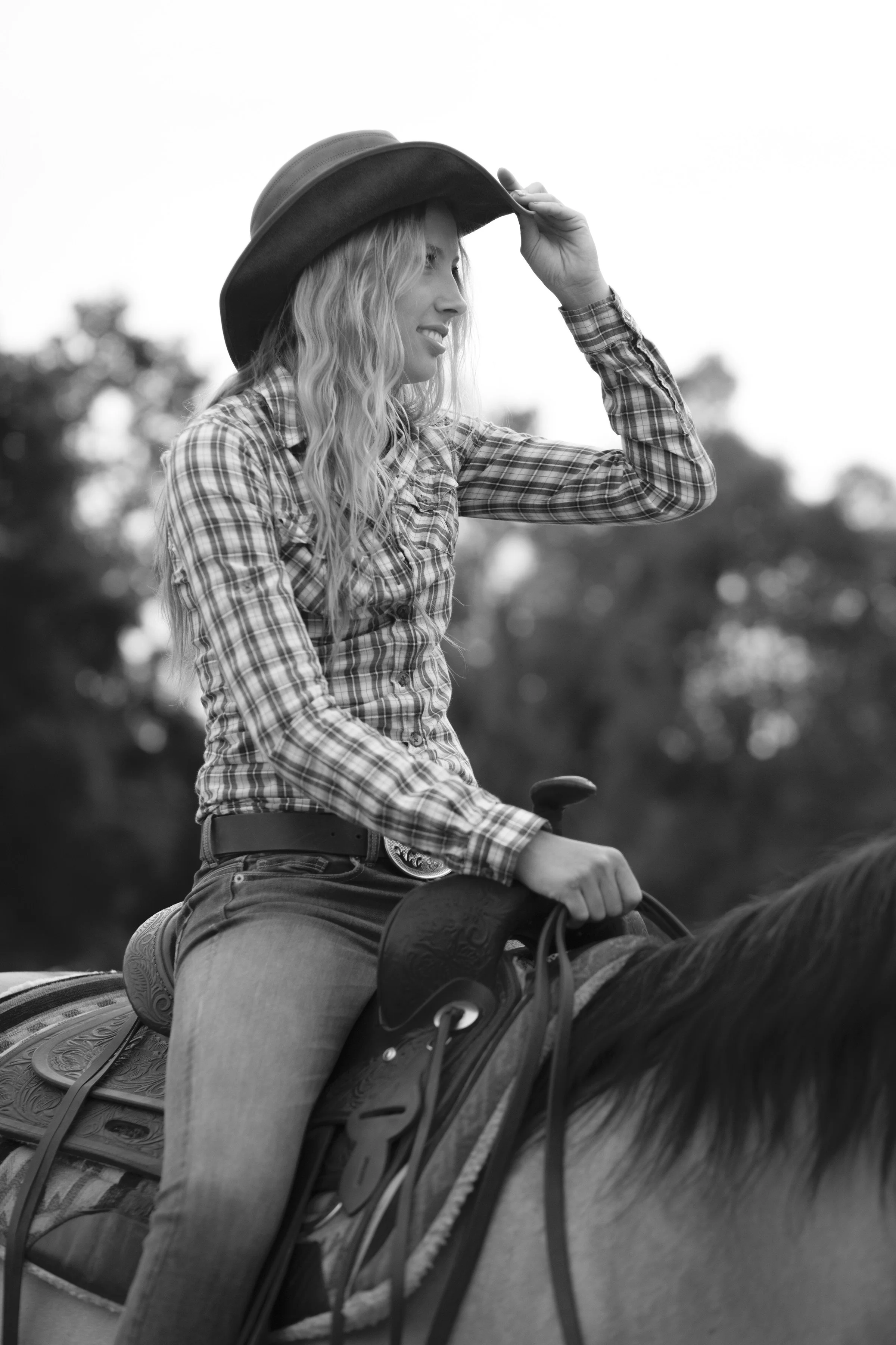 A woman riding a horse, wearing a plaid shirt and a cowboy hat, holding the brim with one hand, outdoors with trees in the background.