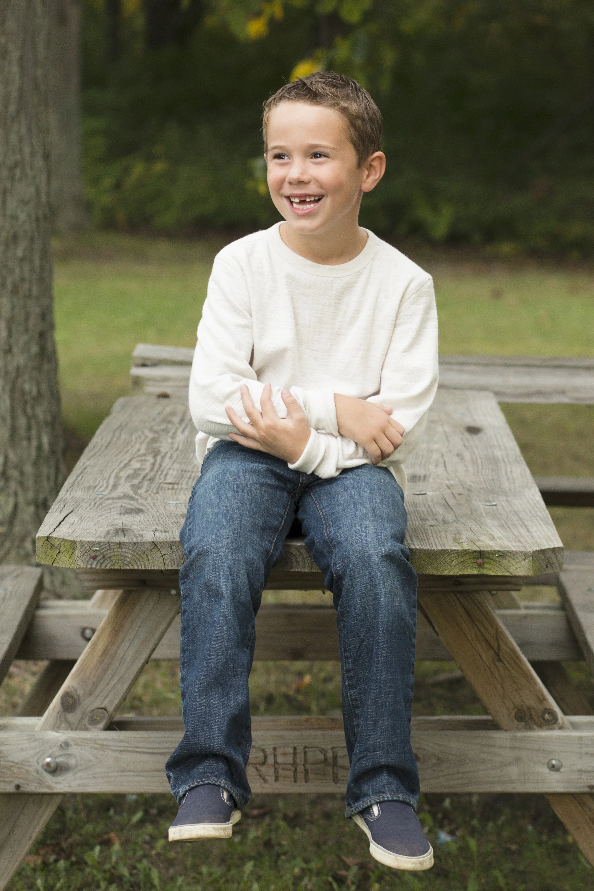 A young boy sitting on a wooden picnic table outdoors, smiling and crossing his arms, with trees and greenery in the background.