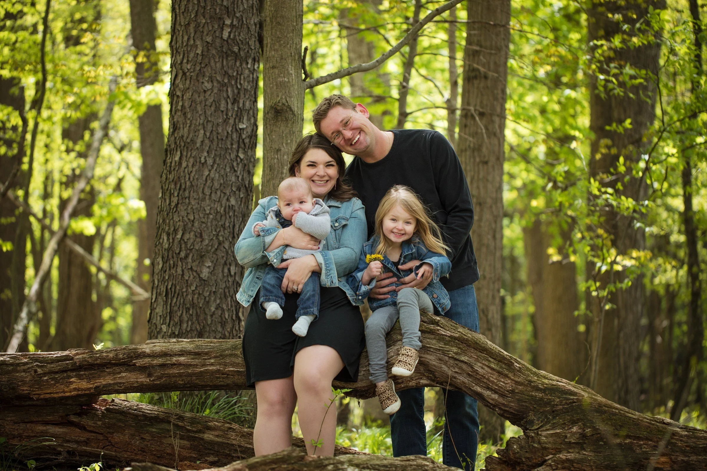 Family of four smiling outdoors in a green forest, sitting on a fallen log.