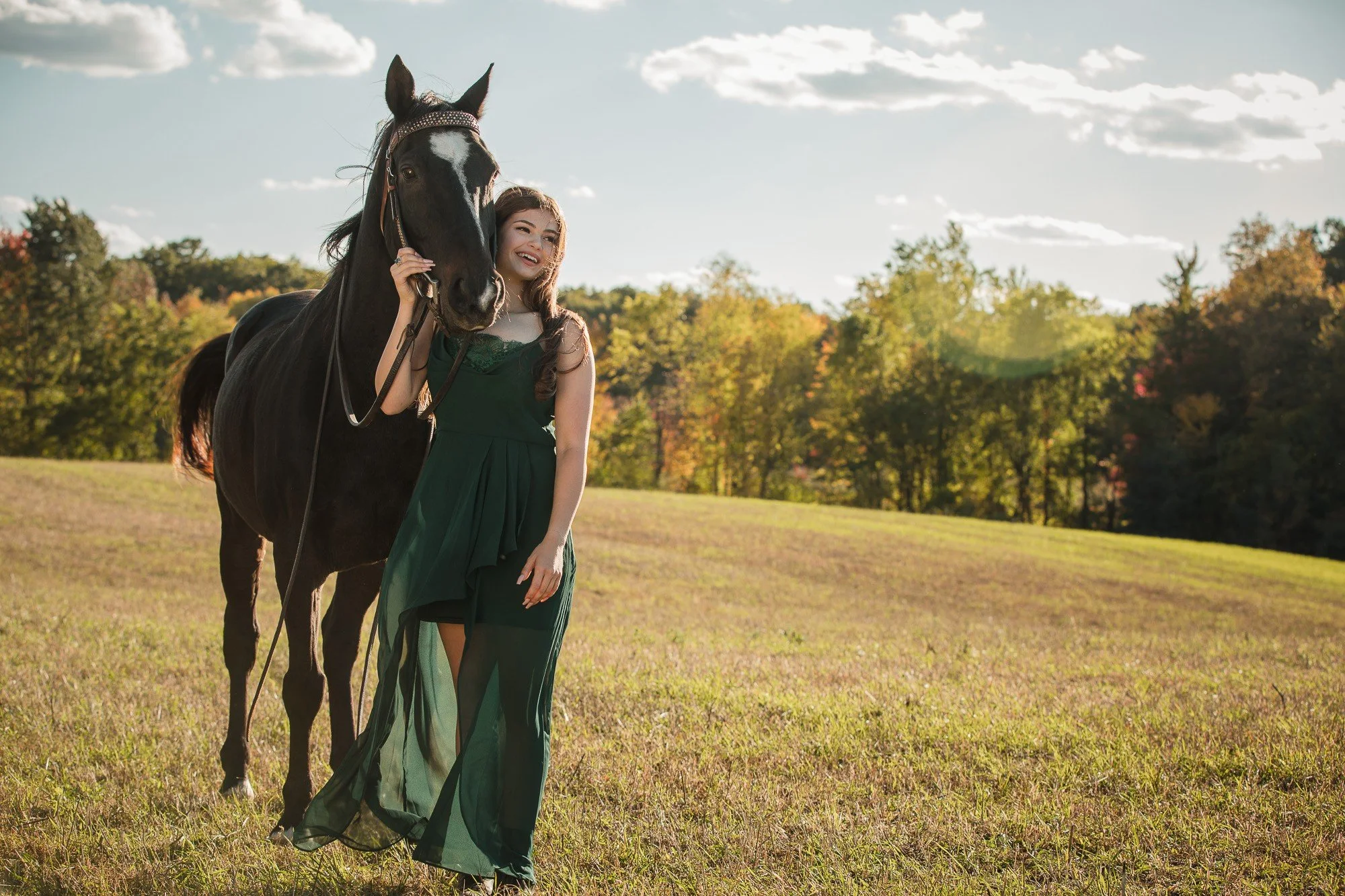 A woman in a green dress standing beside a black horse in an open grassy field with trees and a partly cloudy sky in the background.