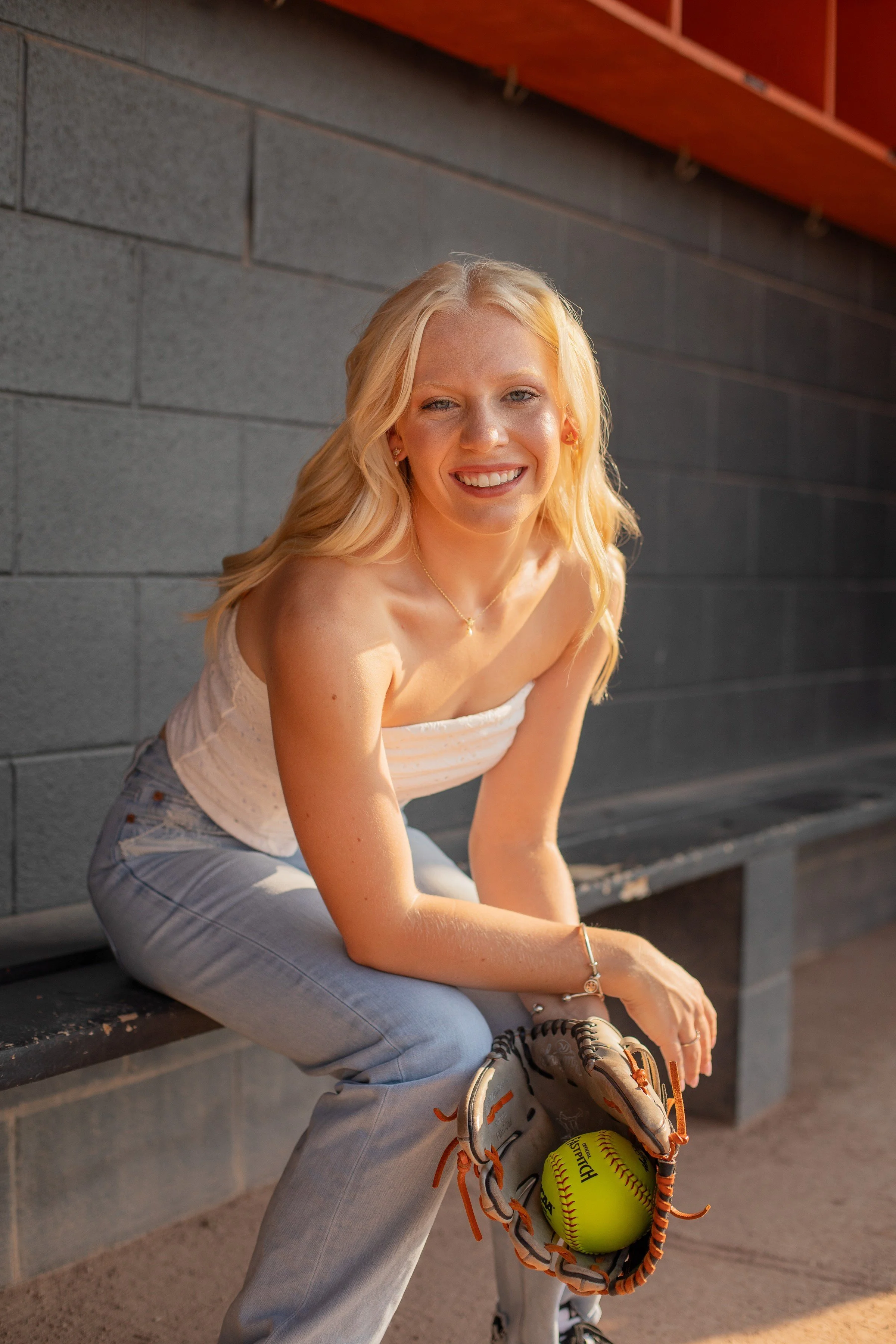 A young woman with blonde hair smiling, sitting on a bench with a softball glove and ball, wearing a white strapless top, grey jeans, and a bracelet.