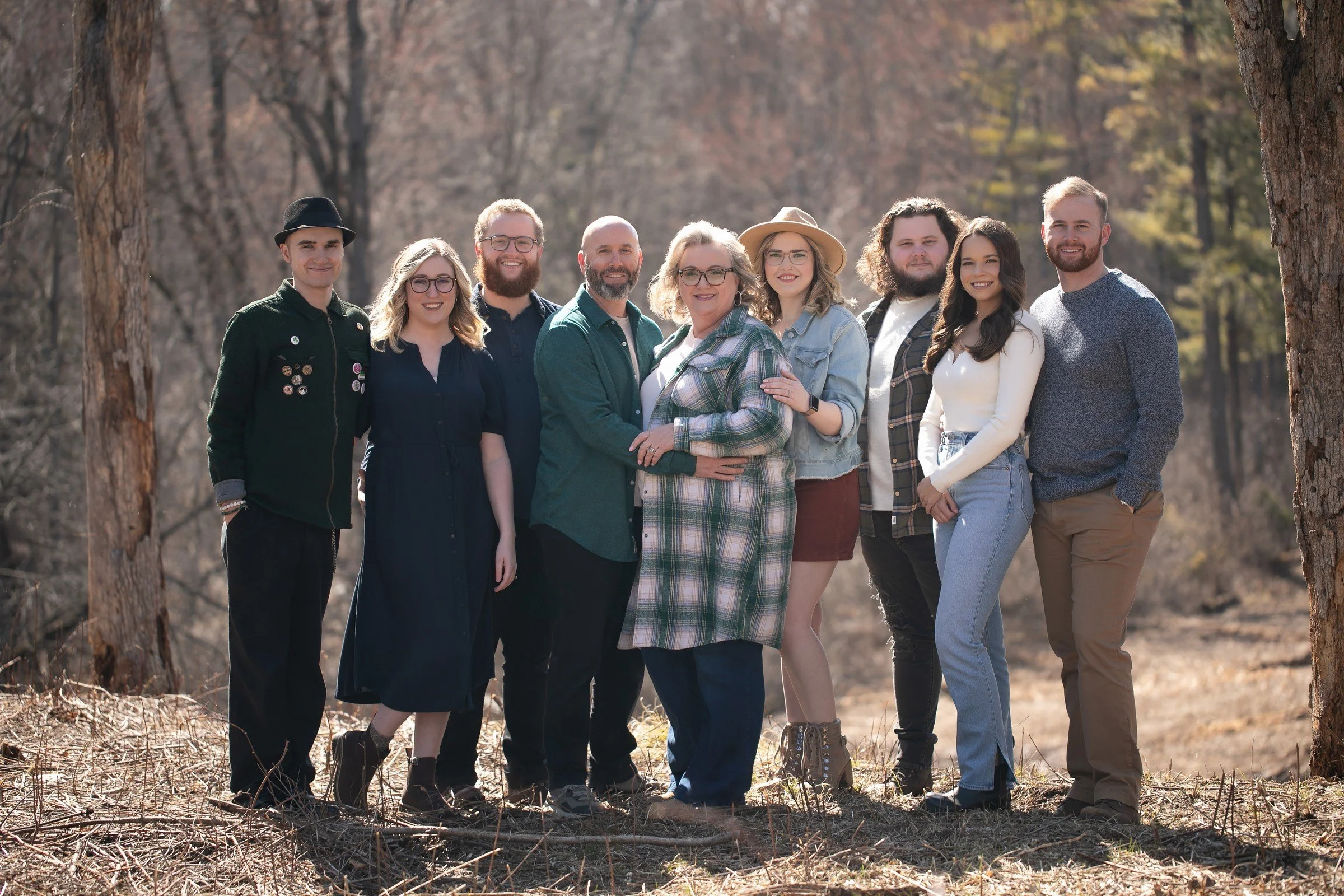 A group of nine people standing together outdoors in a wooded area during daytime, smiling at the camera.
