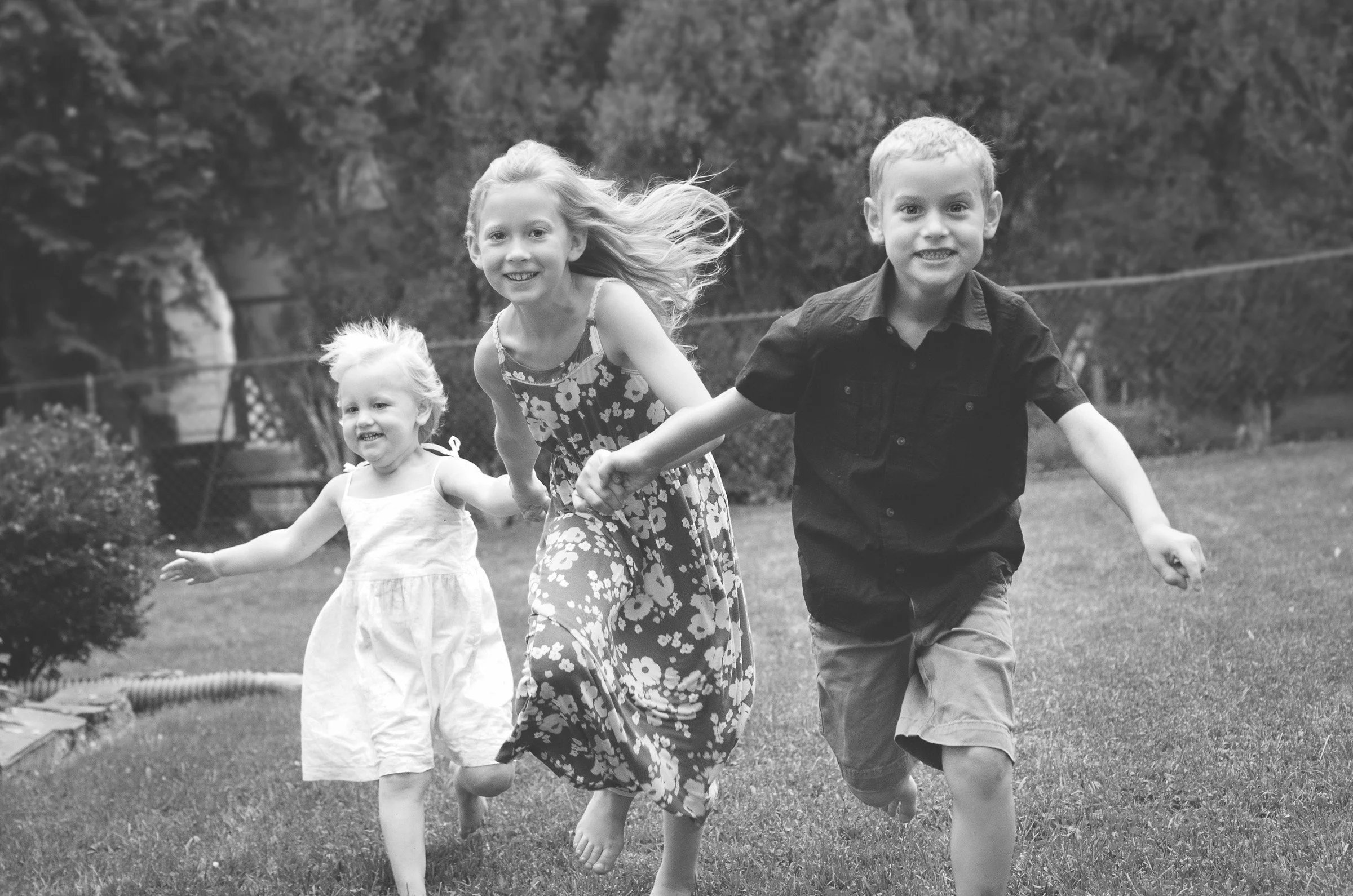 Three children running and holding hands in a grassy yard, smiling and enjoying the outdoors.