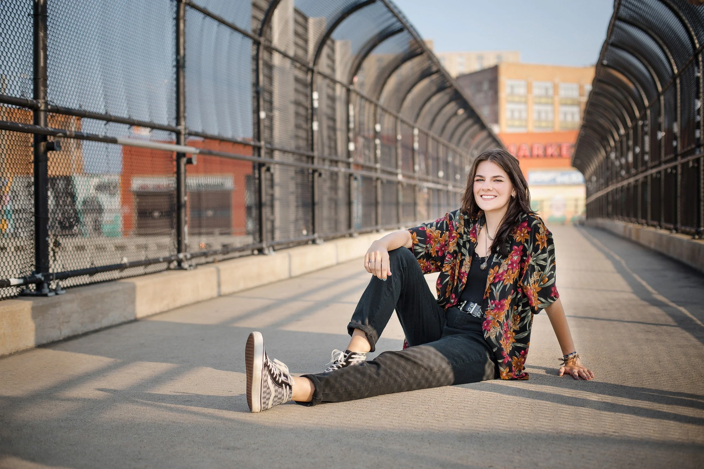 Young woman smiling and sitting on city bridge walkway, wearing black pants, checkered Vans sneakers, floral black shirt, and accessories, with a city skyline and buildings in the background.