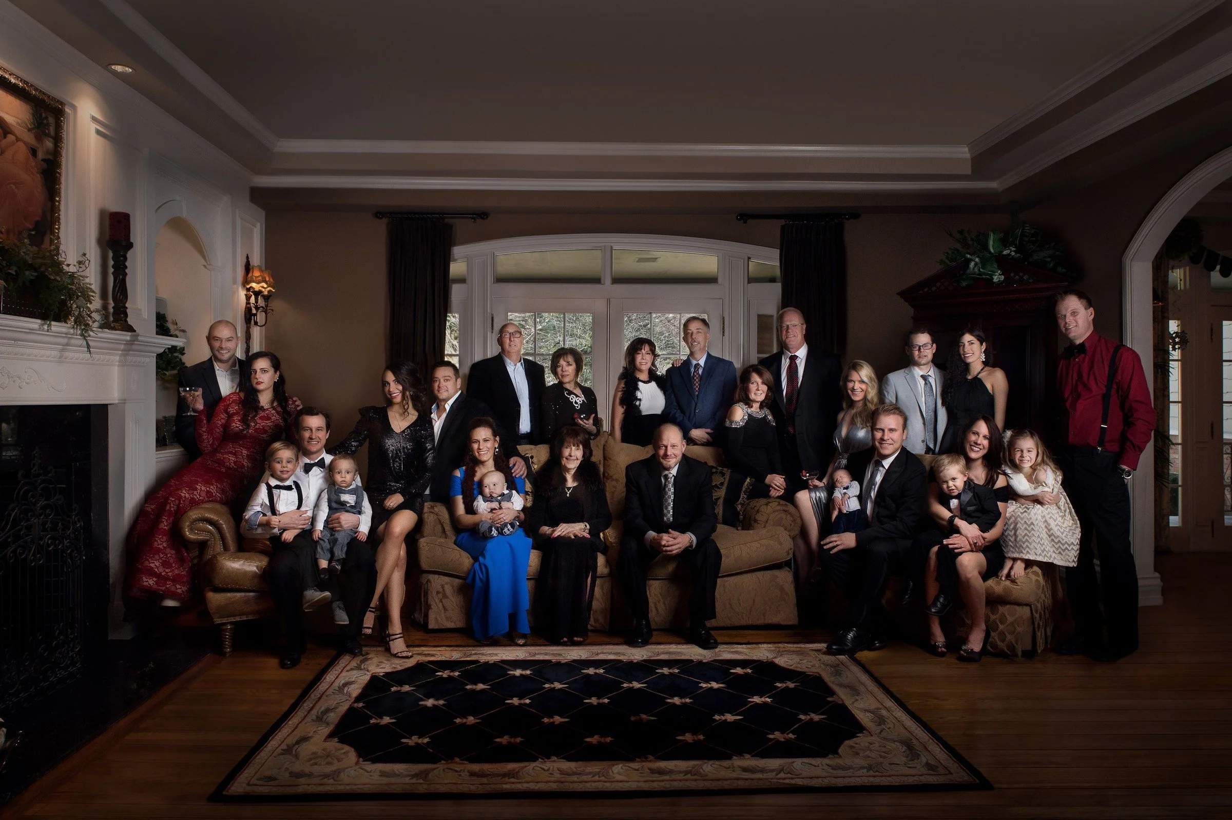 Family gathering in a living room with 23 people, including men, women, and children, dressed in formal and semi-formal attire, sitting and standing around a sofa, with a fireplace, fireplace mantle, and large window with curtains in the background.