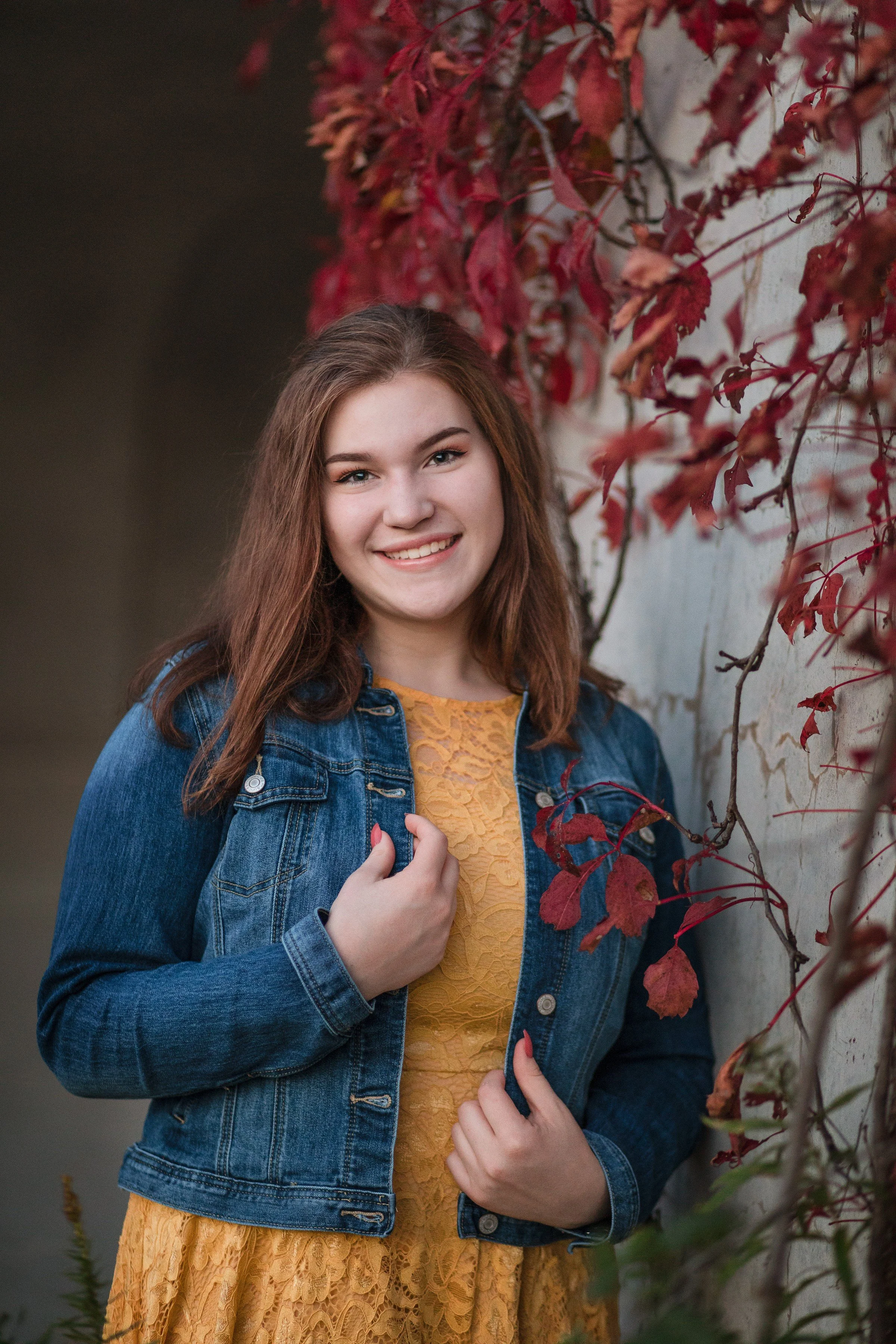 A young woman with shoulder-length brown hair wearing a yellow lace dress and denim jacket standing outdoors next to a white wall with red autumn leaves.