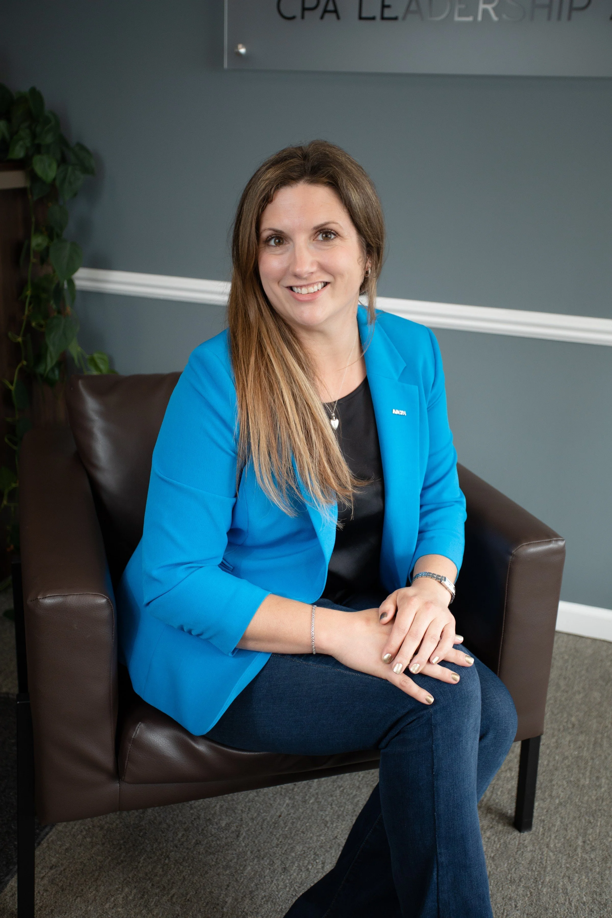 A woman with long brown hair sitting on a dark brown leather chair, wearing a bright blue blazer and smiling. She is in an office setting with a gray wall and a sign that reads 'CPA LEADERSHIP' behind her.