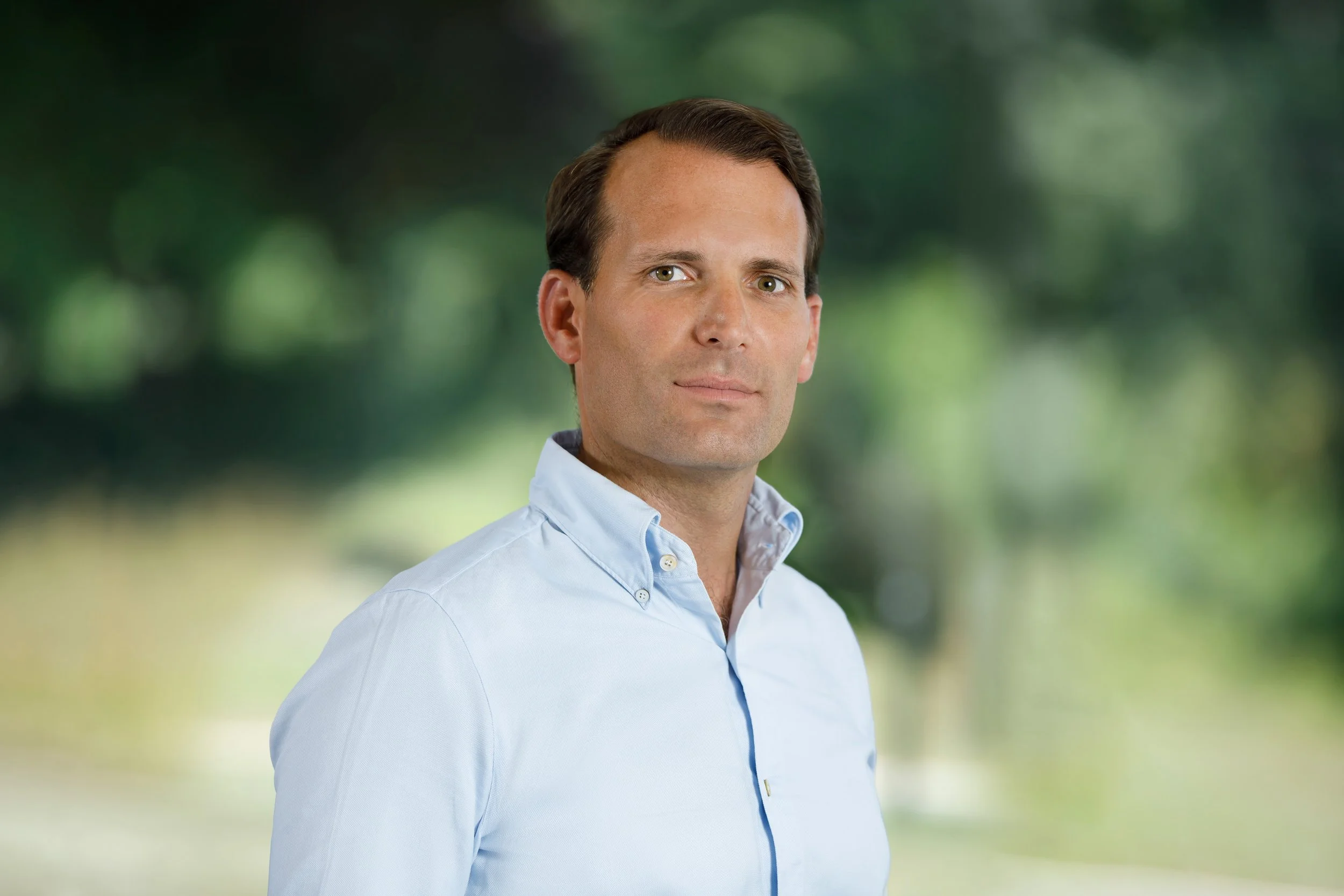 A man with short dark hair wearing a light blue button-down shirt, standing outdoors with a blurred green natural background.