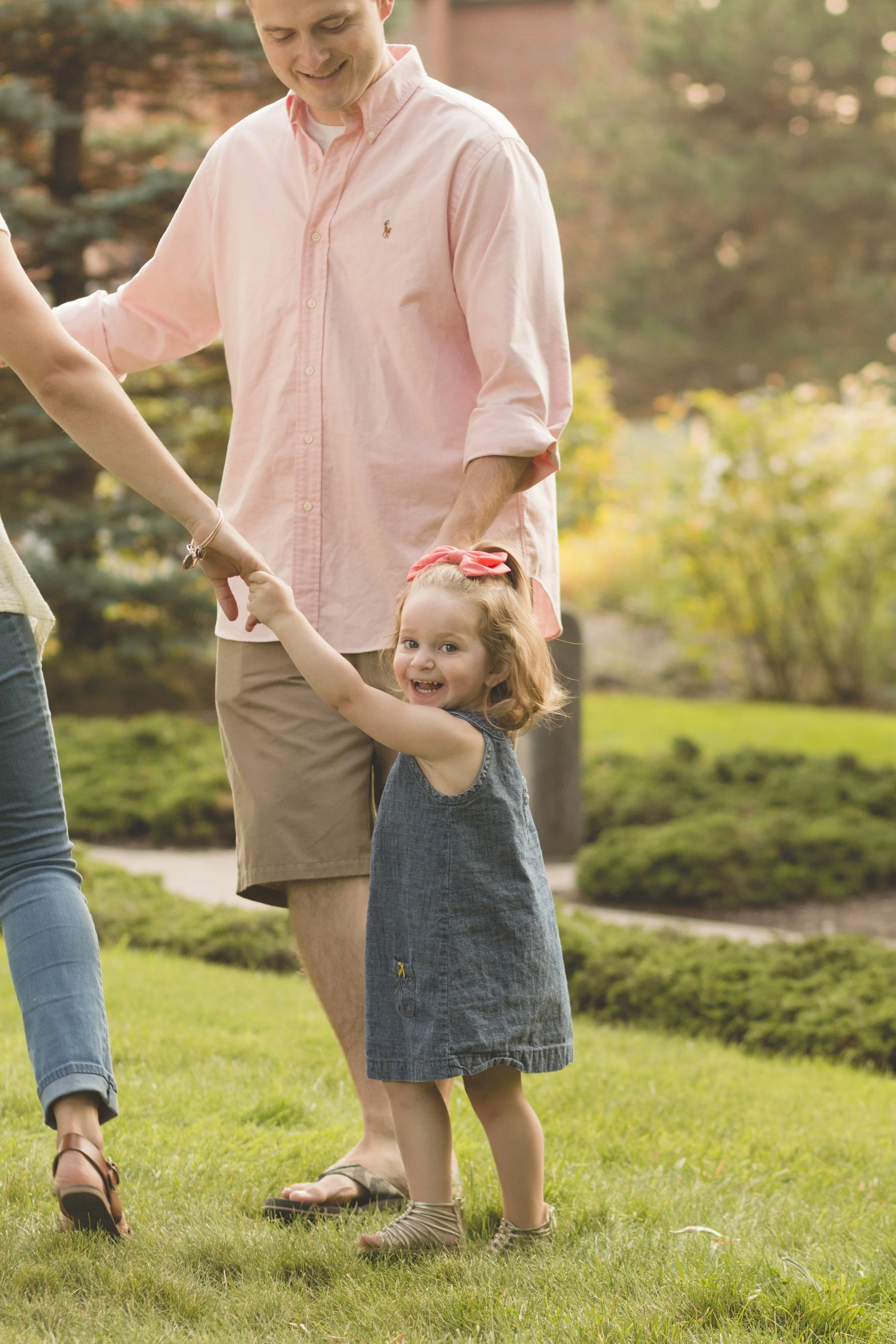 A young girl smiling and holding hands with adults outdoors in a grassy park area.