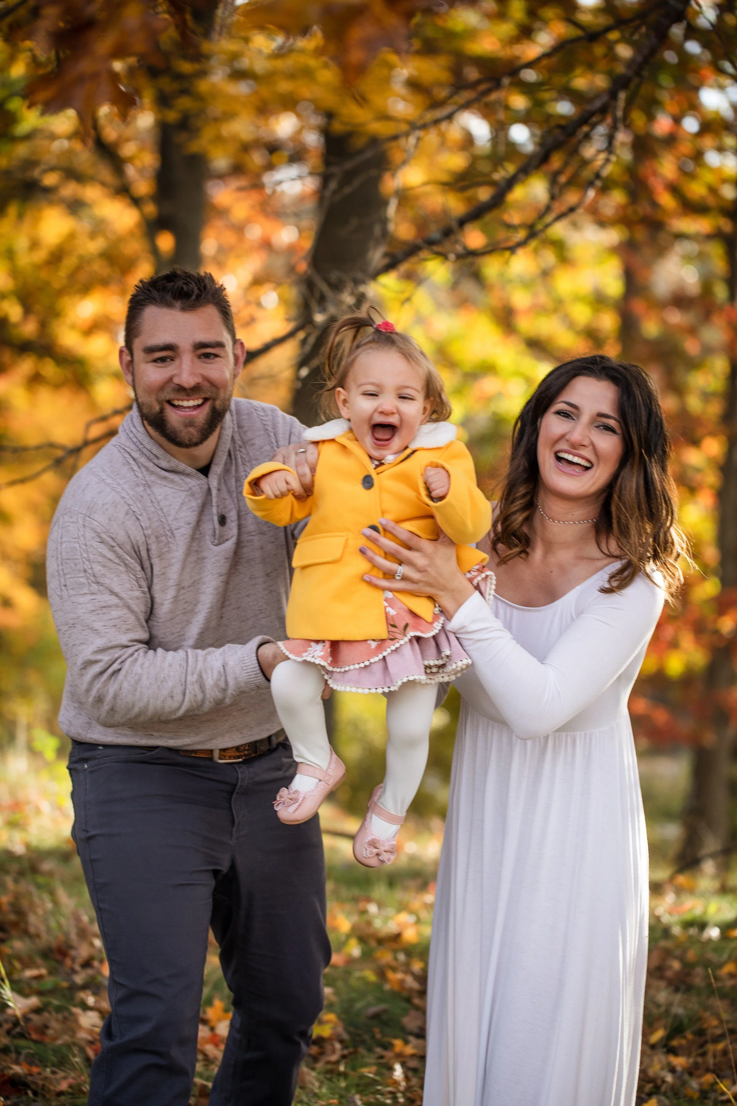 A family of three outside in a park with autumn trees, the father and mother holding up a laughing young girl dressed in a yellow coat.