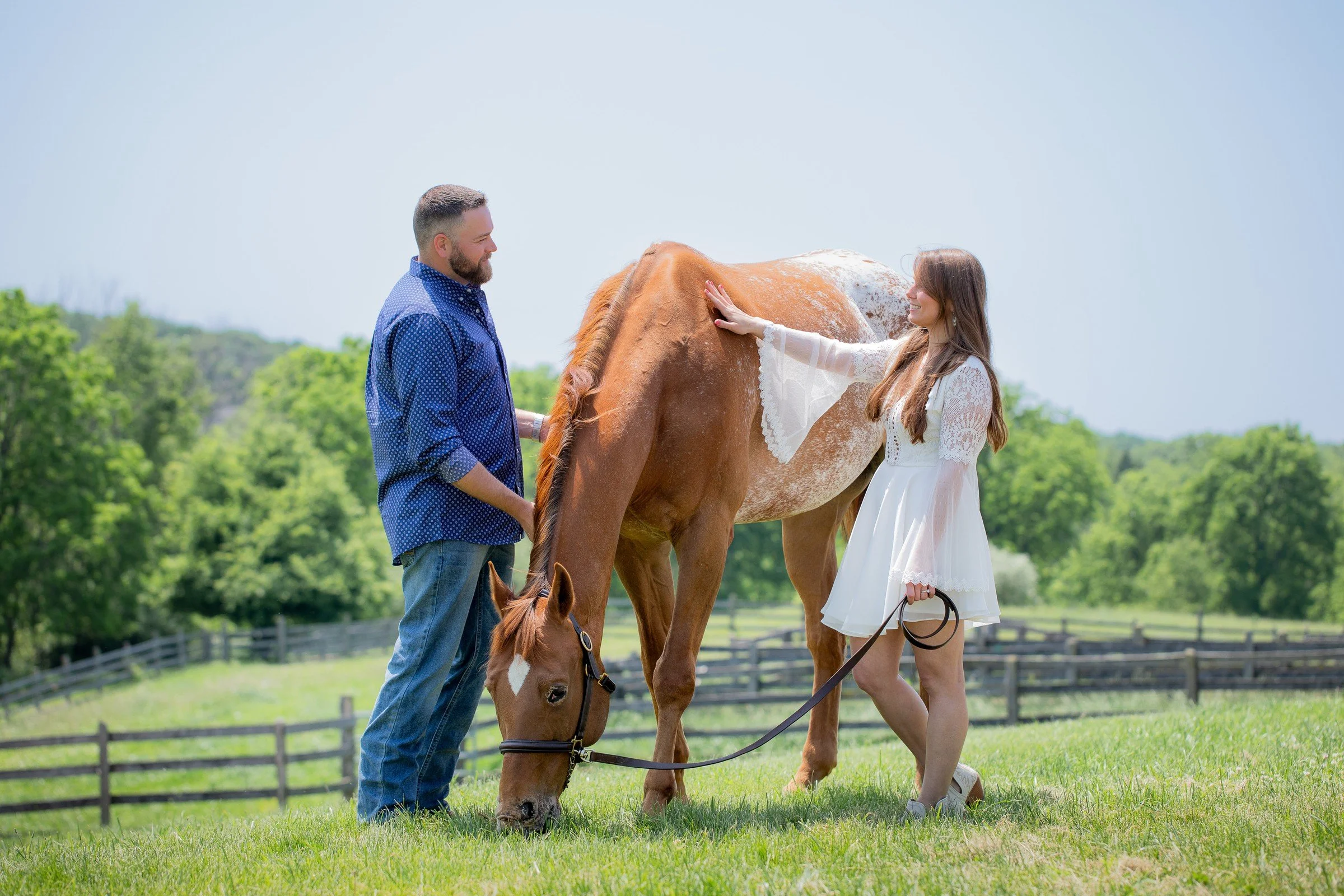 A woman in a white dress petting a brown horse while a man stands nearby holding the horse's reins on a green field with trees and a fence in the background.