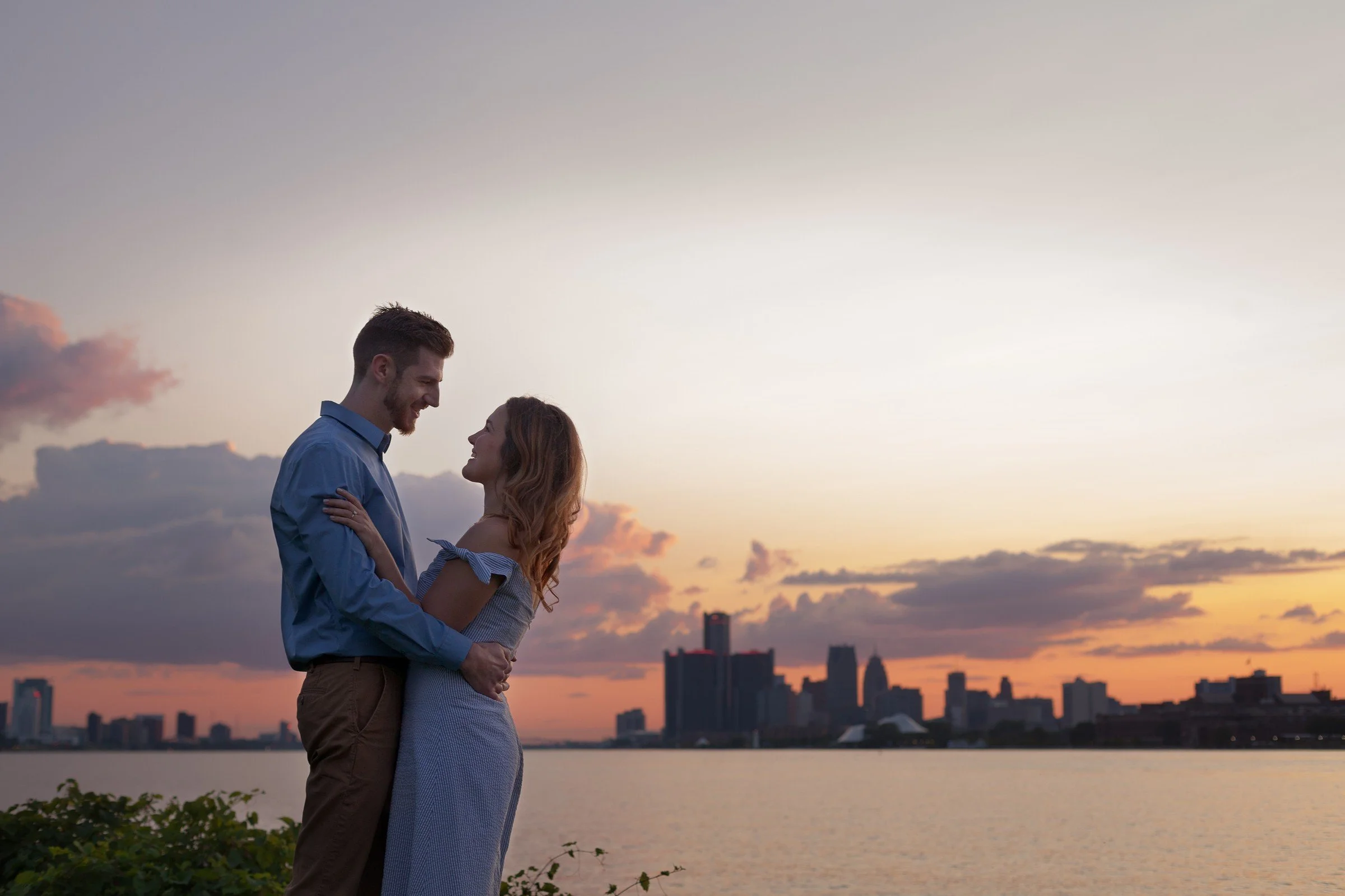 A couple embraces each other at sunset near a river, with a city skyline in the background.
