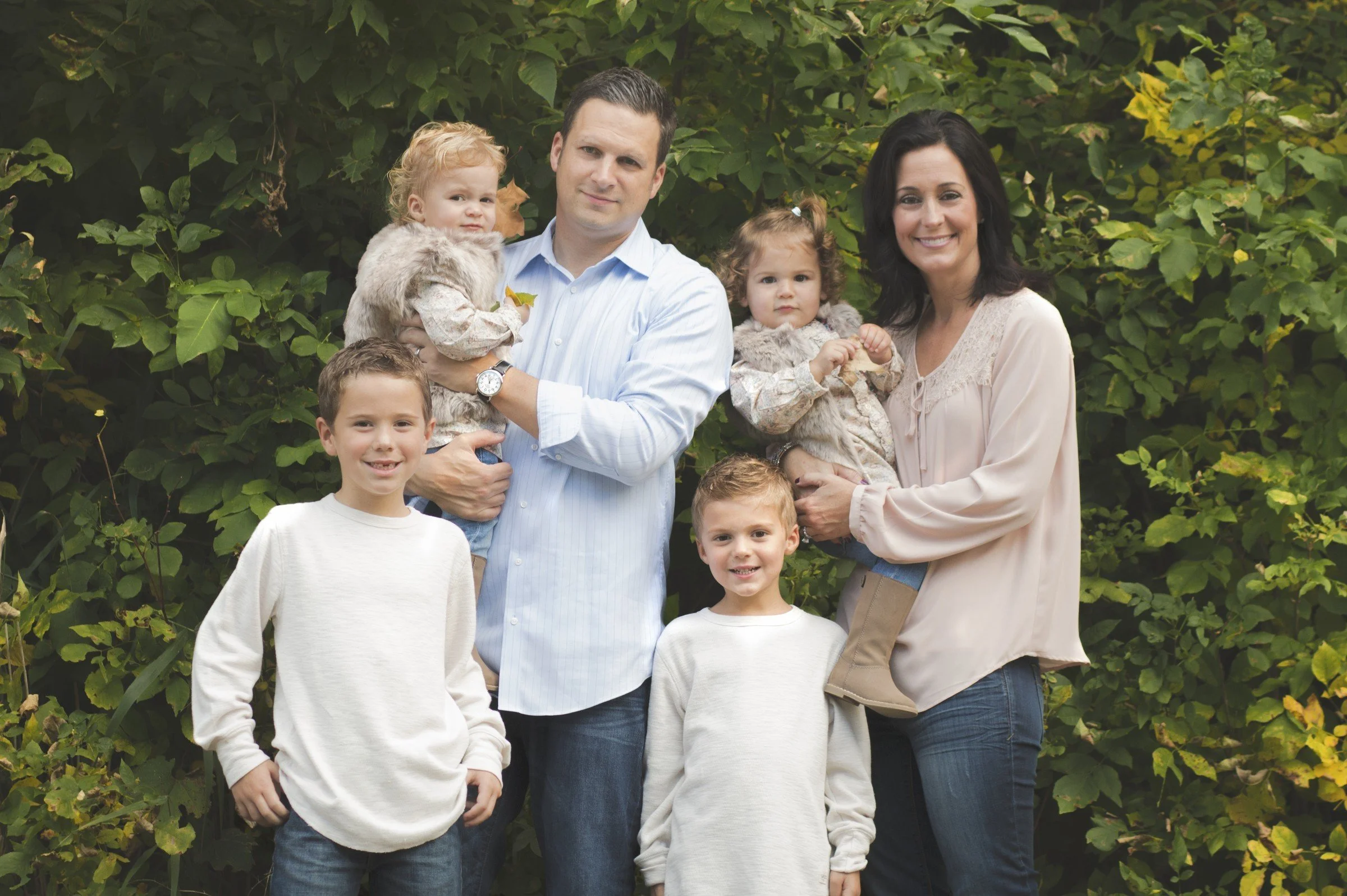 A family of seven posing outdoors in front of green foliage, including two adults and five children, some being held by the adults.