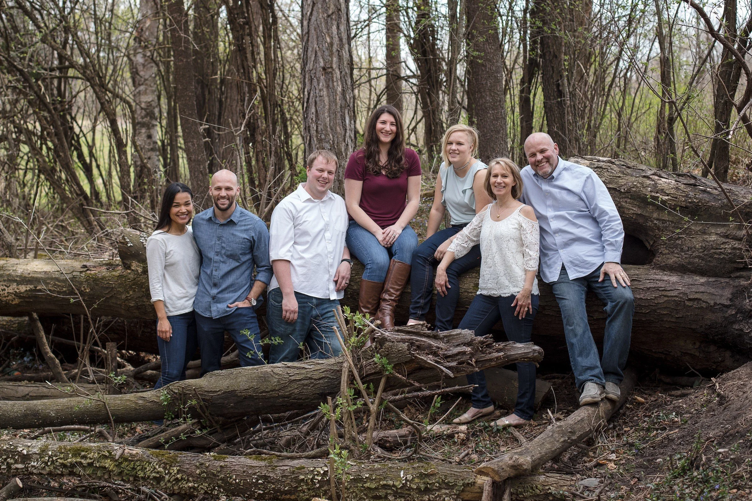 A group of seven people posing on a fallen tree in a forest, smiling, with bare trees and branches around them.