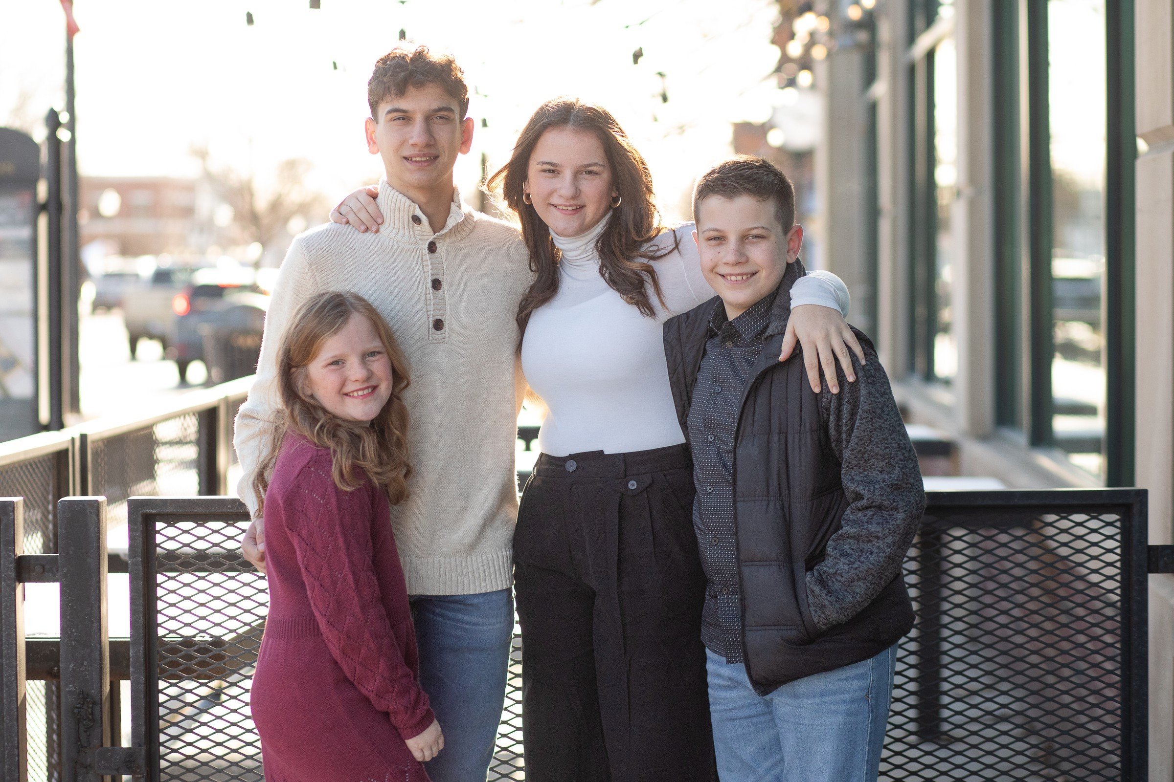 Group of five people standing outside, smiling, with sunlight in the background. Three children and two adults, two boys and a girl. Everyone has their arms around each other, standing near a railing.