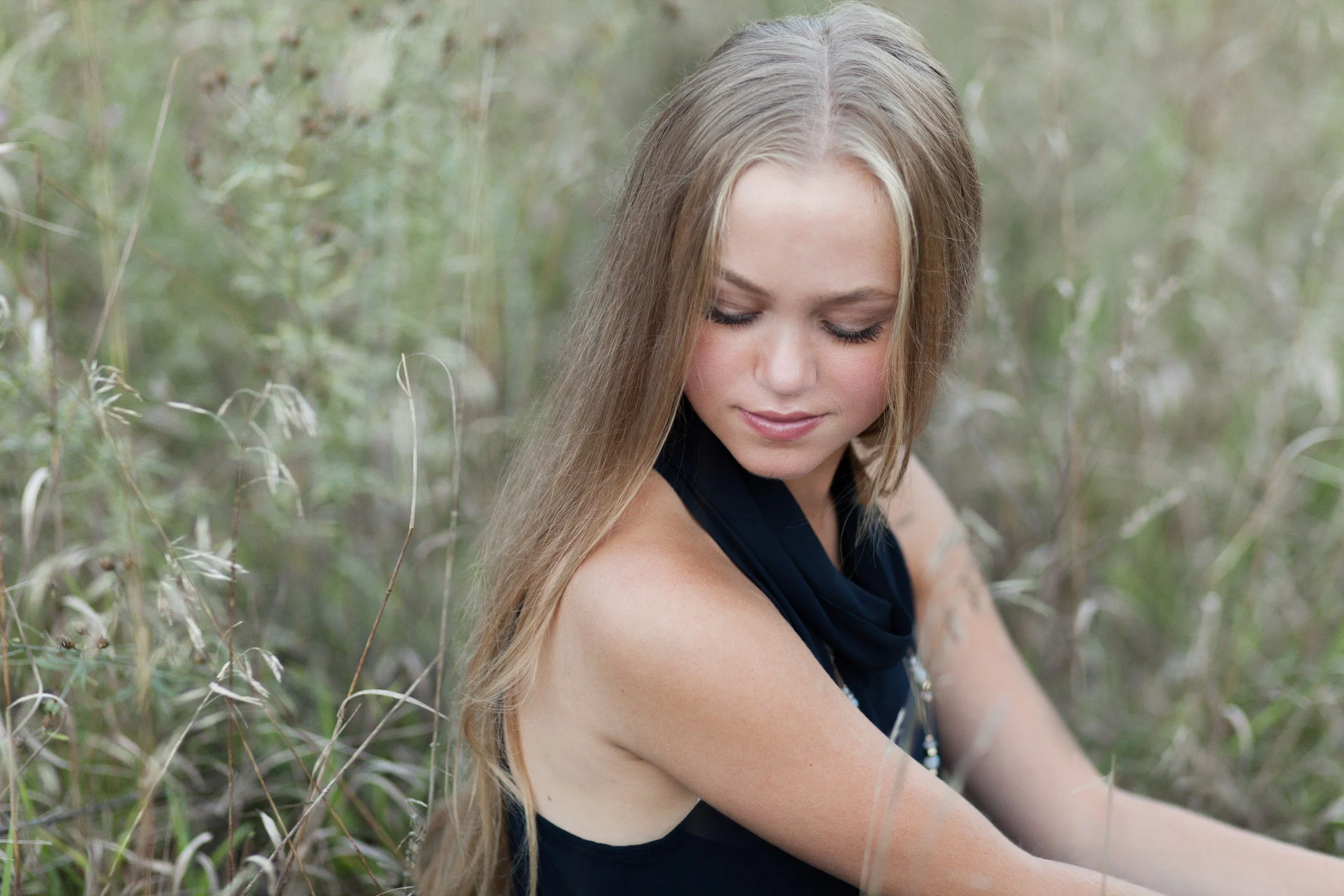 A young woman with long blonde hair sitting outdoors in tall grass, wearing a sleeveless black top and a blue scarf, looking down with a peaceful expression.