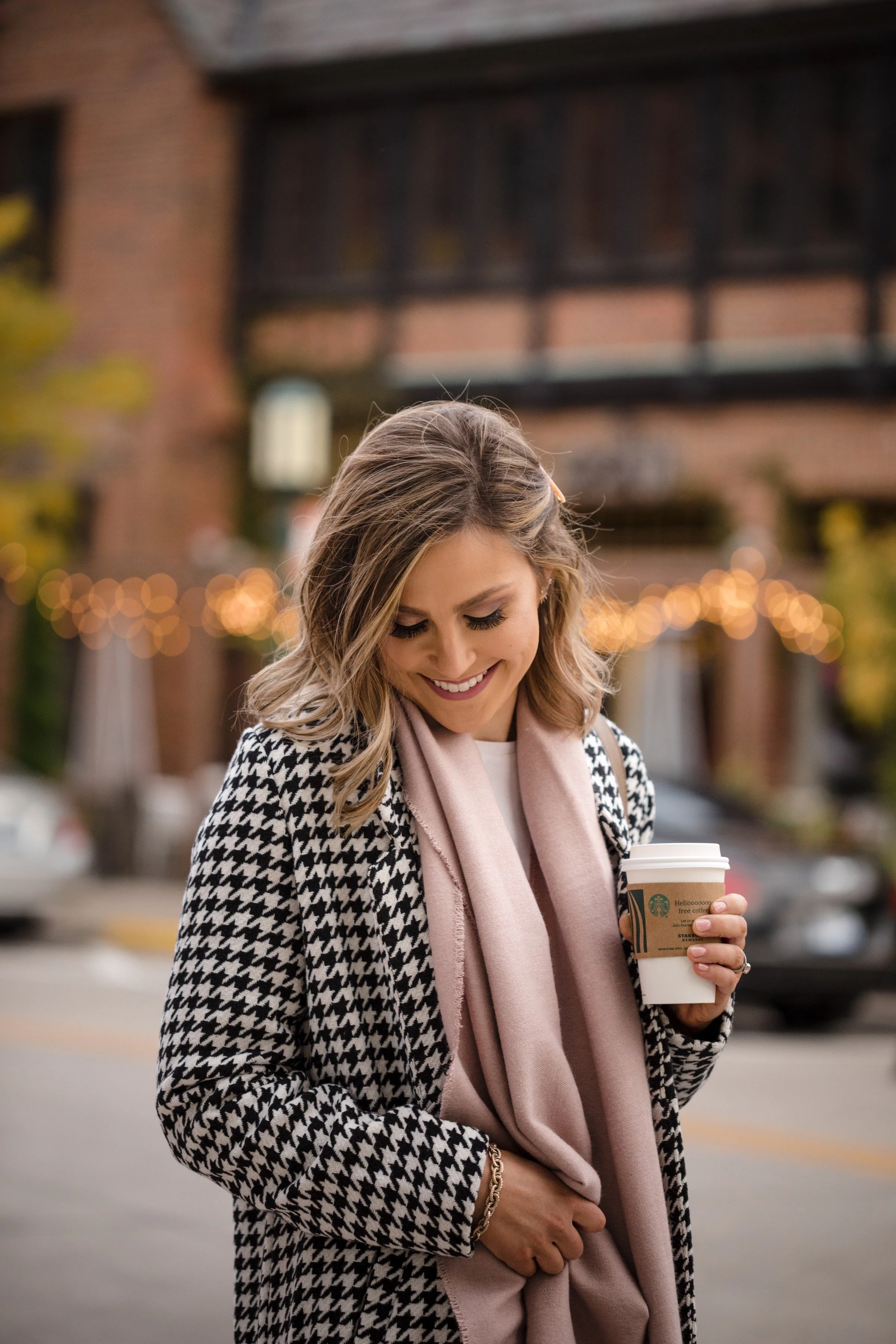 A smiling woman with shoulder-length wavy blonde hair, wearing a black and white houndstooth coat and a light pink scarf, holding a to-go coffee cup in an outdoor setting with blurred background lights and a brick building.