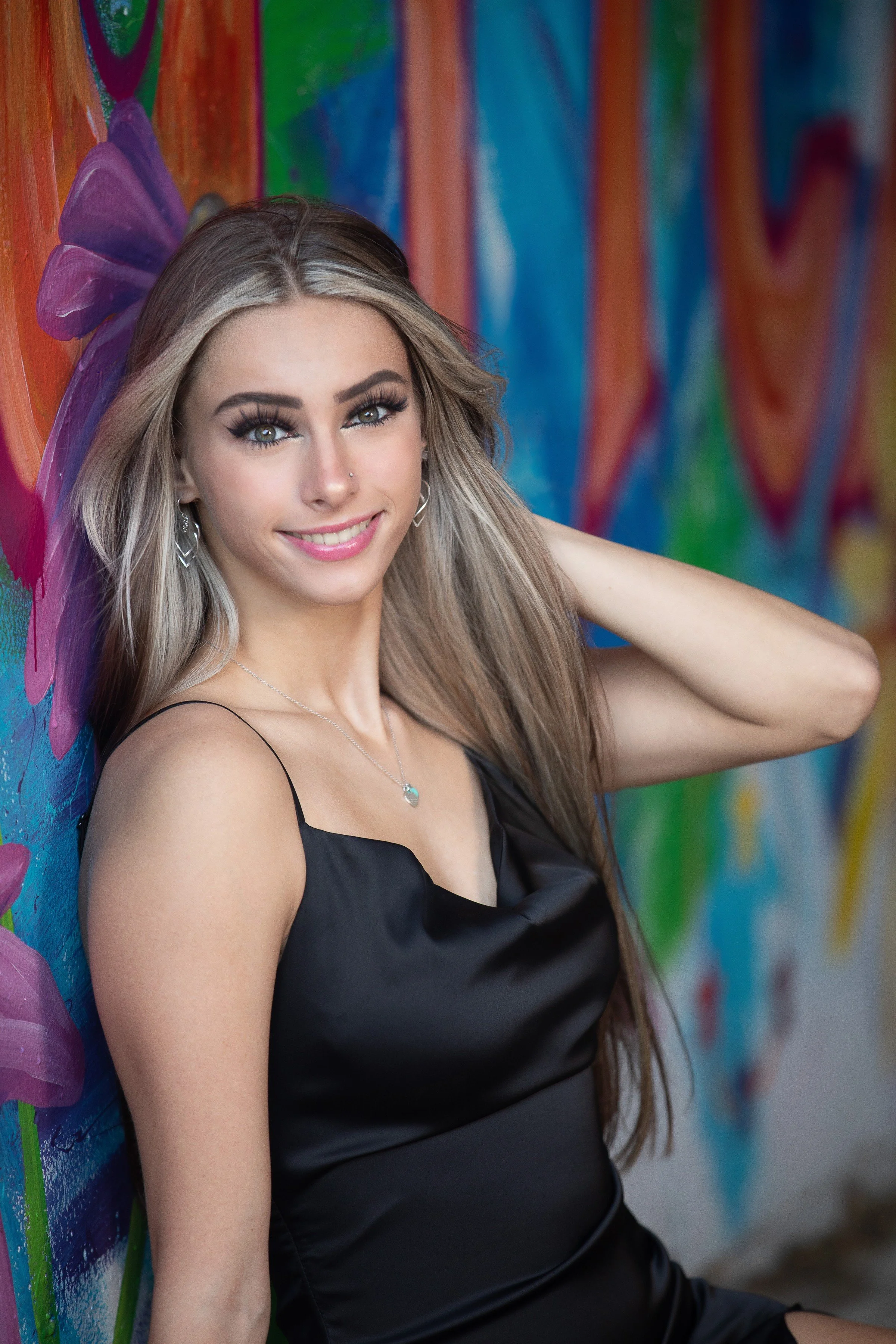 A young woman with long blonde hair, wearing a black satin dress, smiling and posing with her left hand behind her head against a colorful graffiti wall.
