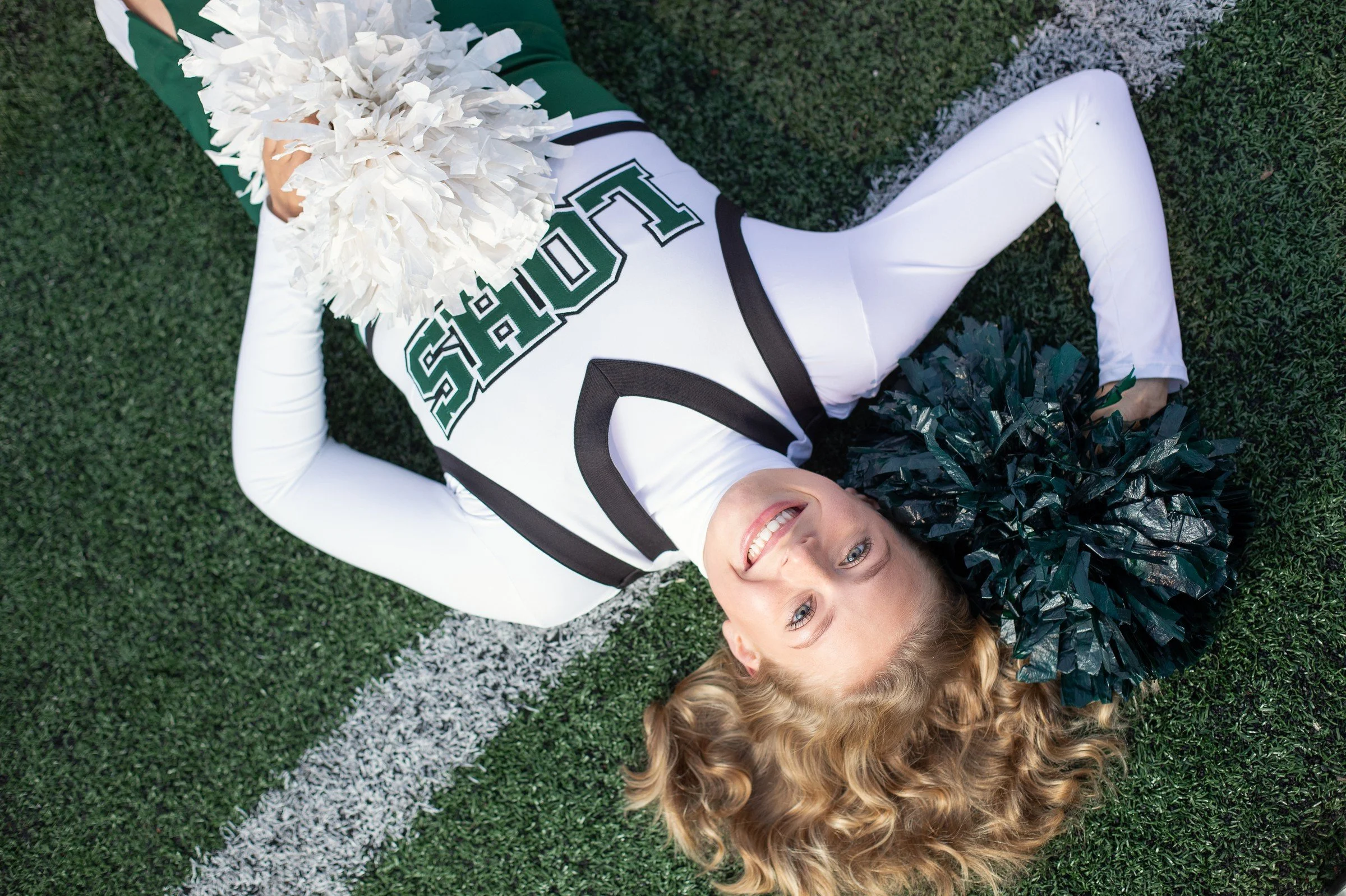 A cheerleader with blonde curly hair lying on a football field, smiling, holding pom-poms, wearing a white and green uniform with the letters 'SPARTS'.
