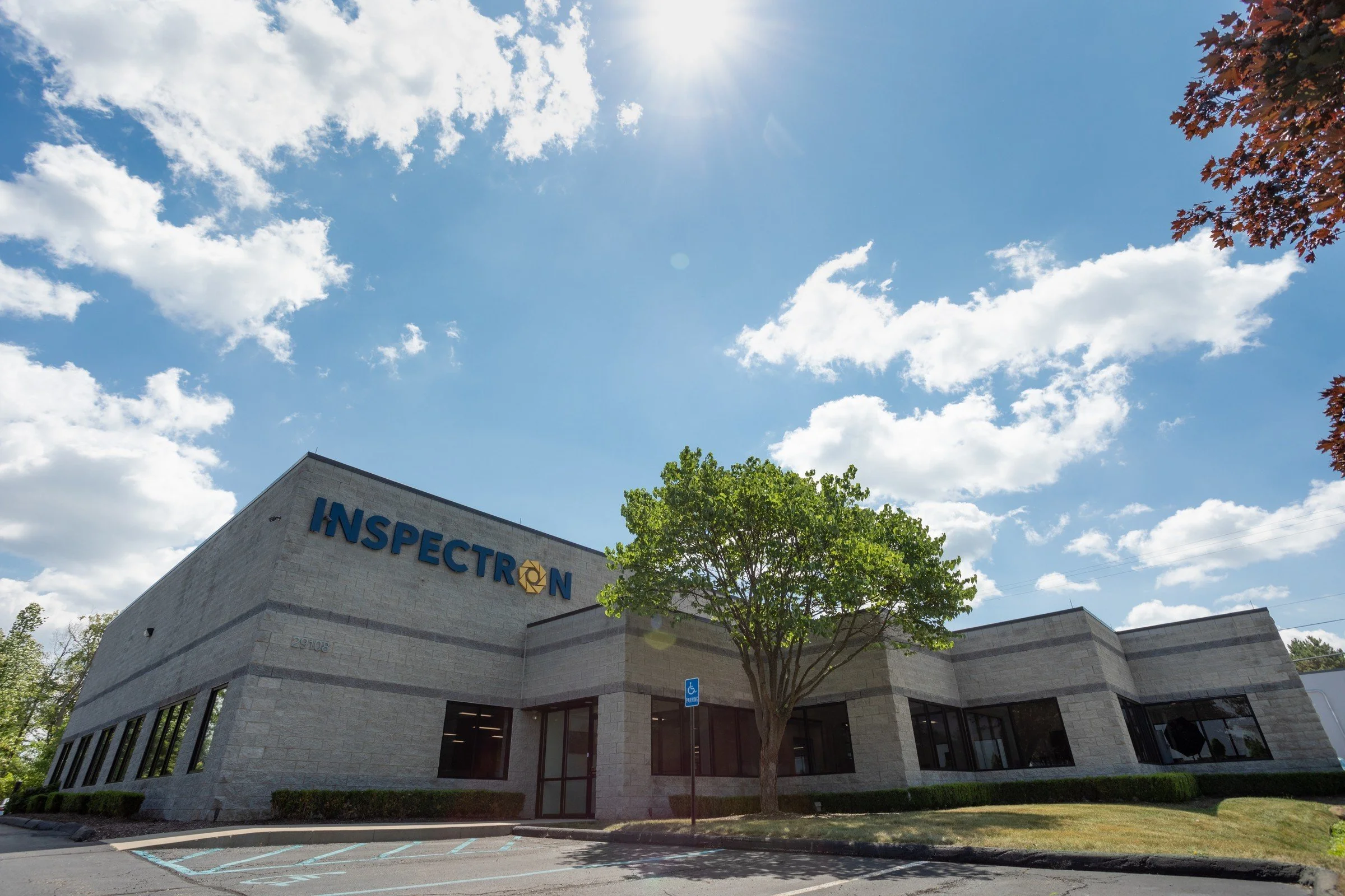 Exterior view of a modern office building with the sign 'INSPECTION' on the front, a parking lot, a handicap parking sign, a tree, and a partly cloudy blue sky.