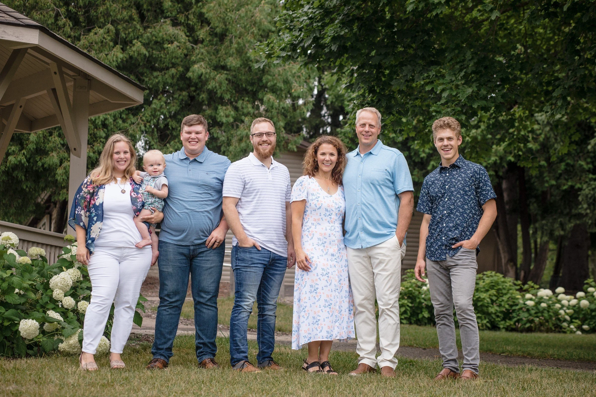 Family photo of seven people, including a baby, standing outdoors on grass in front of trees and bushes, smiling and posing for the camera.