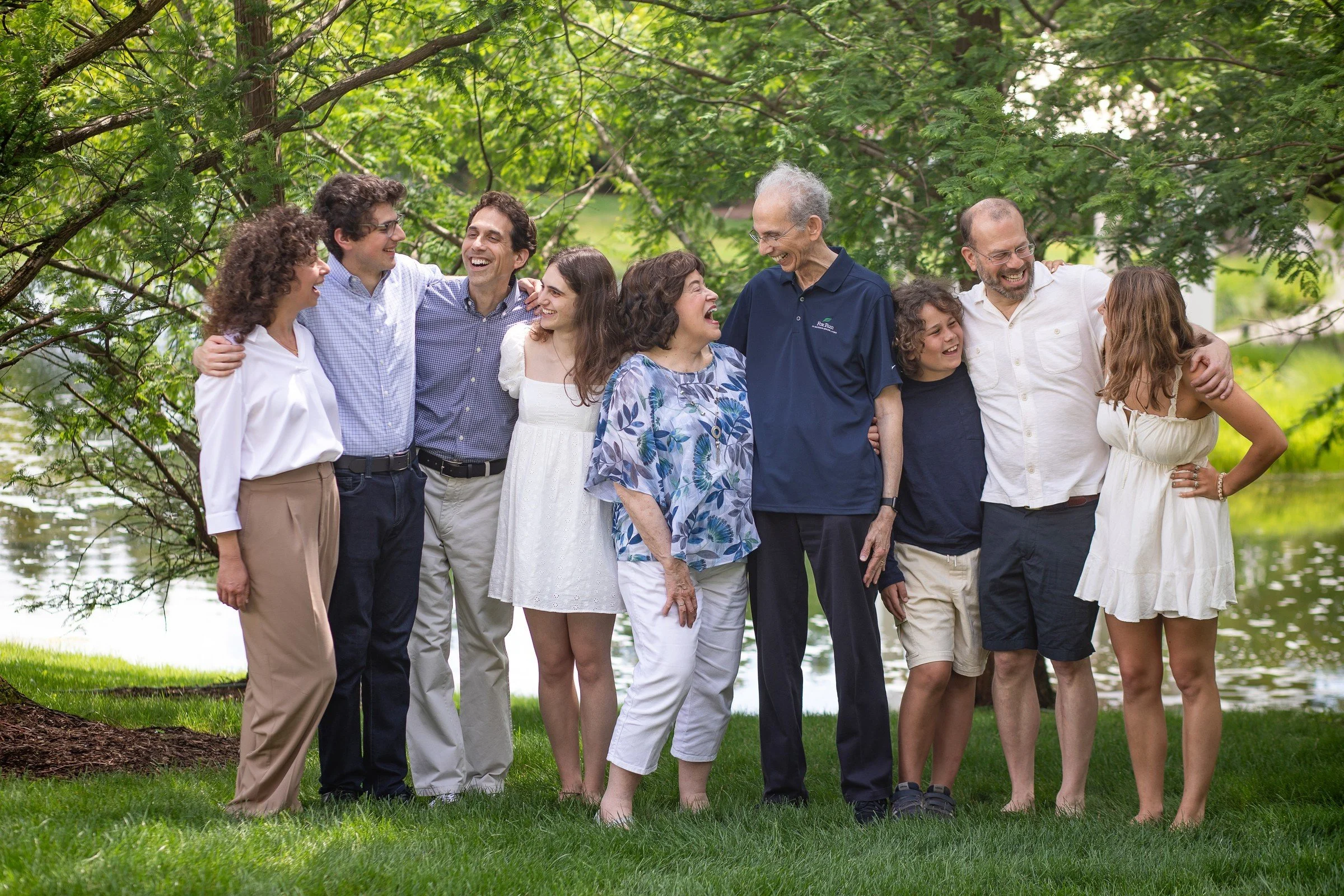 Group of nine people, adults and children, standing outdoors under a tree near water, smiling and laughing, embracing each other.