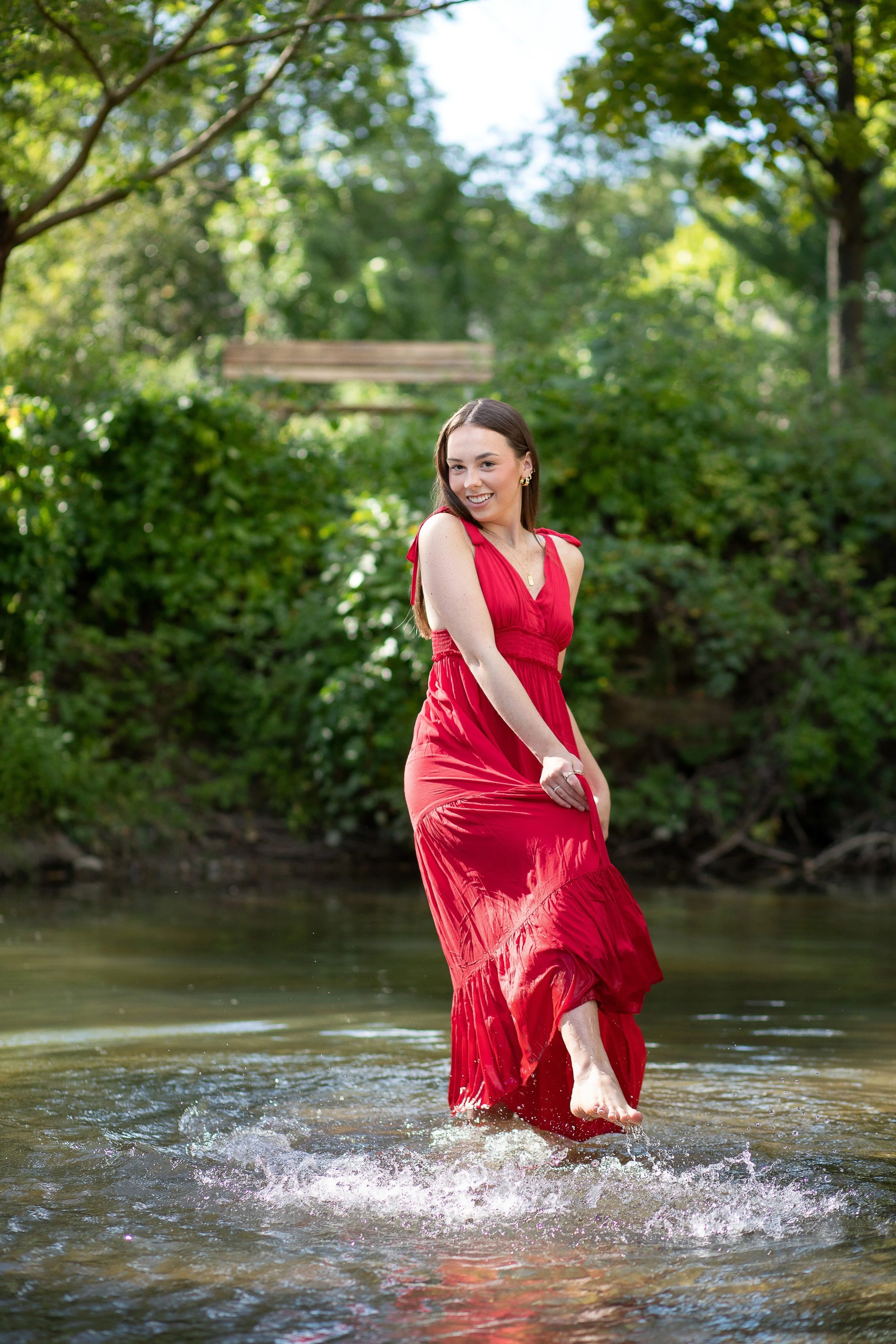A woman in a red dress smiling and walking barefoot in a shallow creek surrounded by lush green trees.