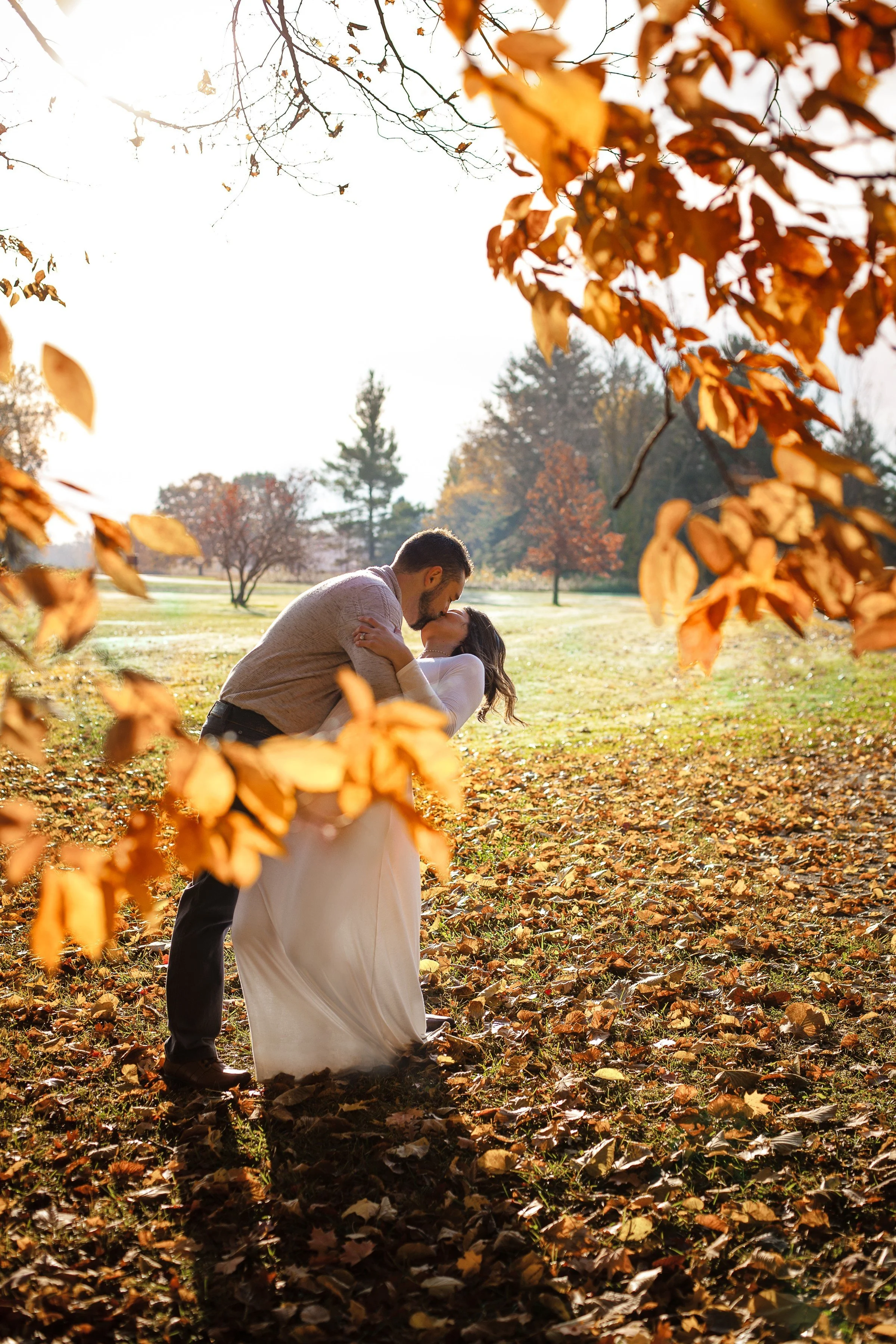 A couple in wedding attire kissing in a park with autumn leaves on the ground and trees with fall foliage in the background.