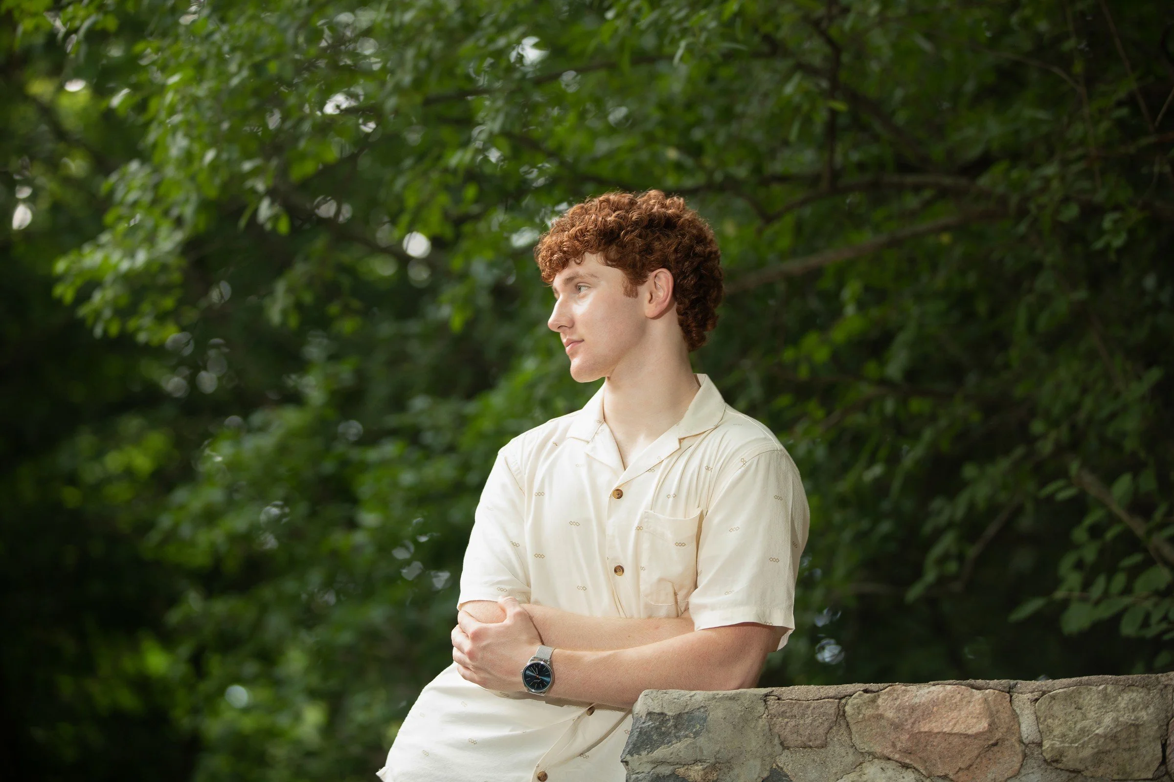 A young man with curly red hair wearing a light-colored short-sleeve shirt and a watch, leaning on a stone wall and looking to the side in a lush, green outdoor setting.