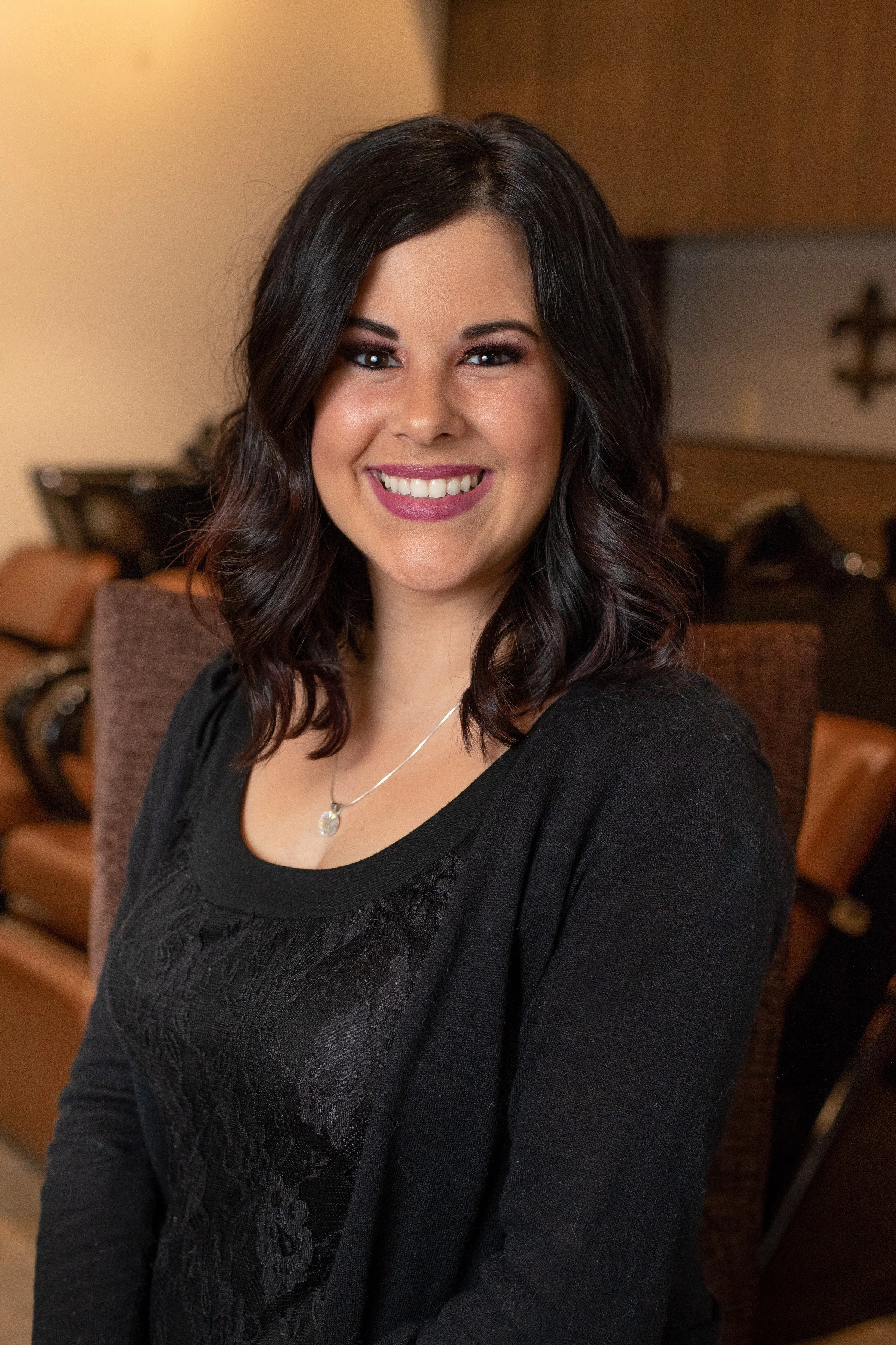 A woman with dark, wavy hair and a bright smile, wearing a black top and a silver necklace, sitting indoors with a warm background.