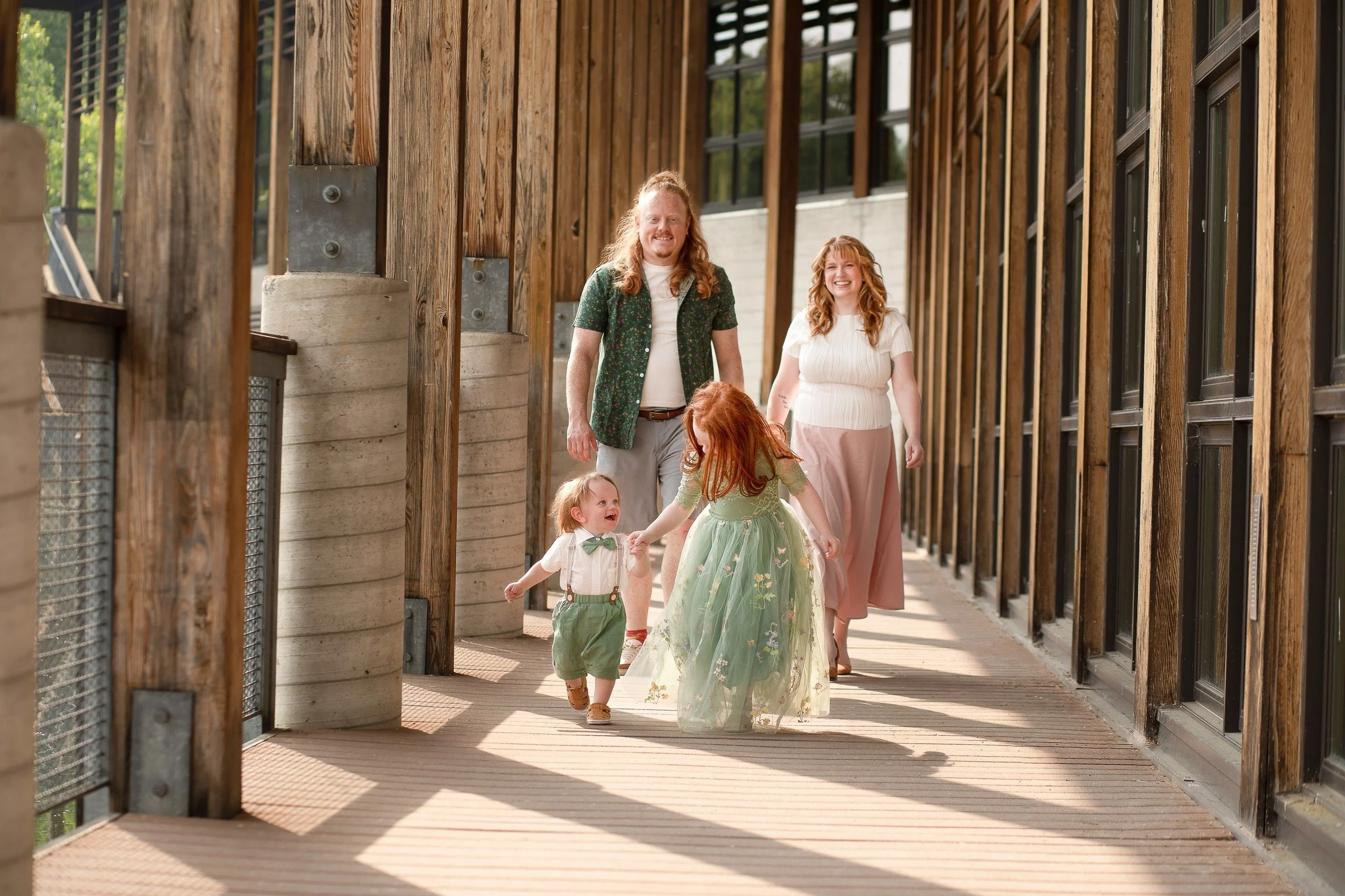 A family walking down a wooden corridor outdoors, with two children and two adults, all smiling and enjoying a sunny day.