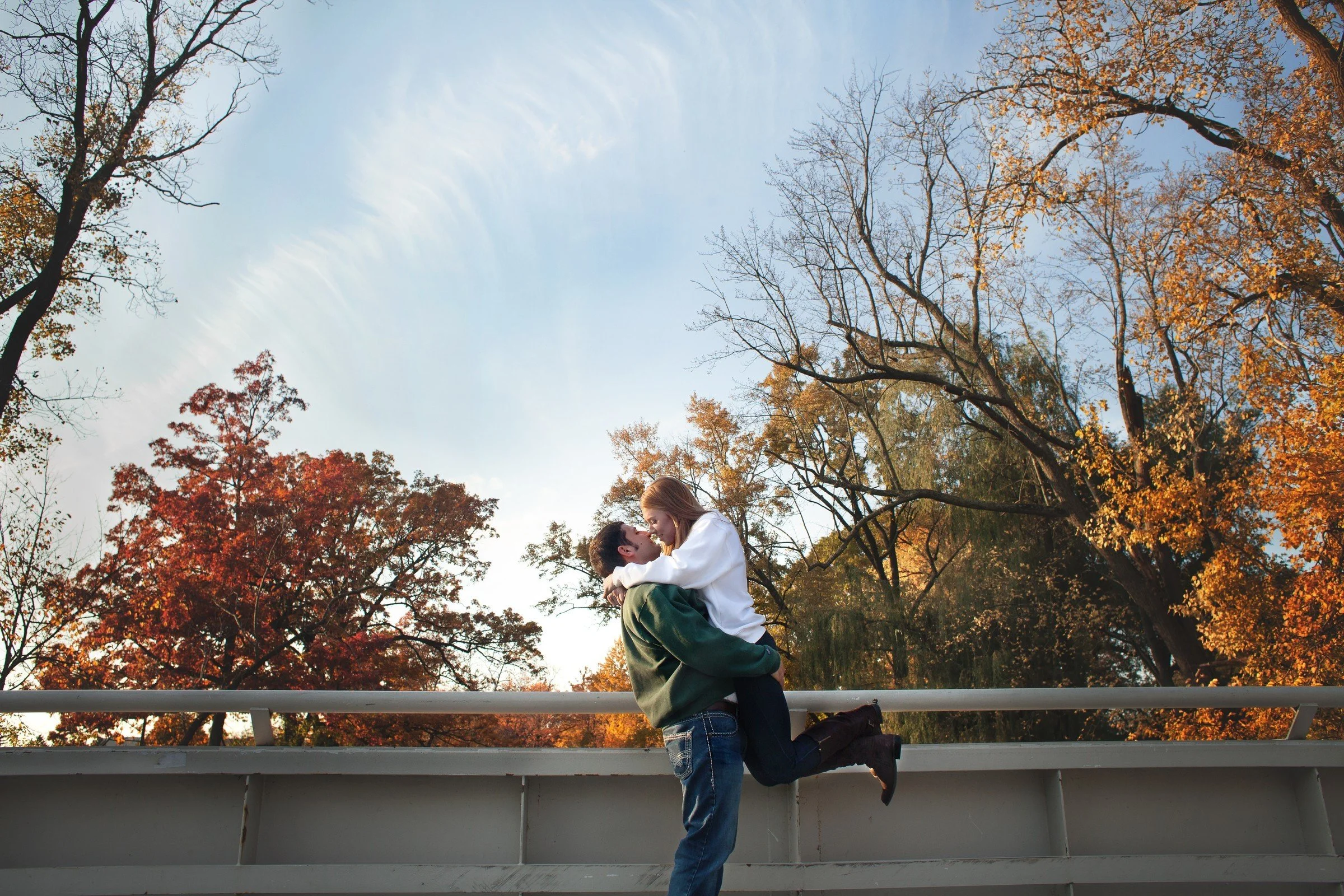 A couple embracing on a bridge during autumn, surrounded by trees with orange and yellow leaves, with a blue sky overhead.