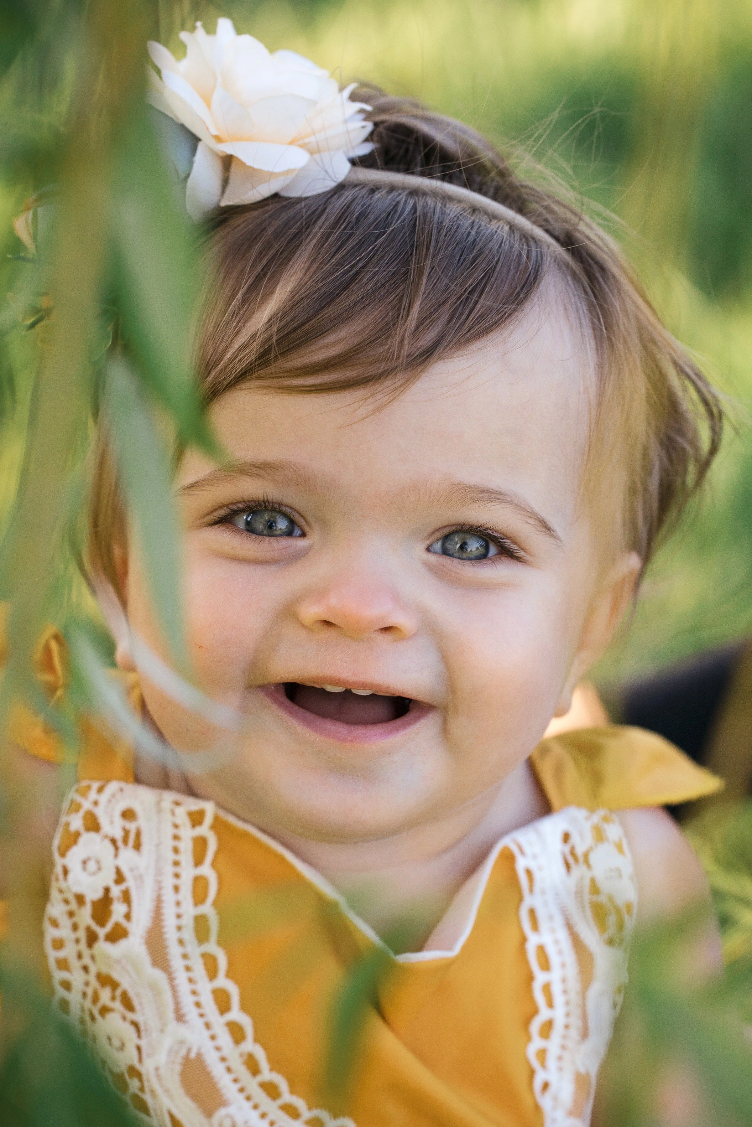 Close-up of a smiling toddler girl with blue eyes, wearing a yellow dress with lace details, outdoors with greenery and a white flower headband.