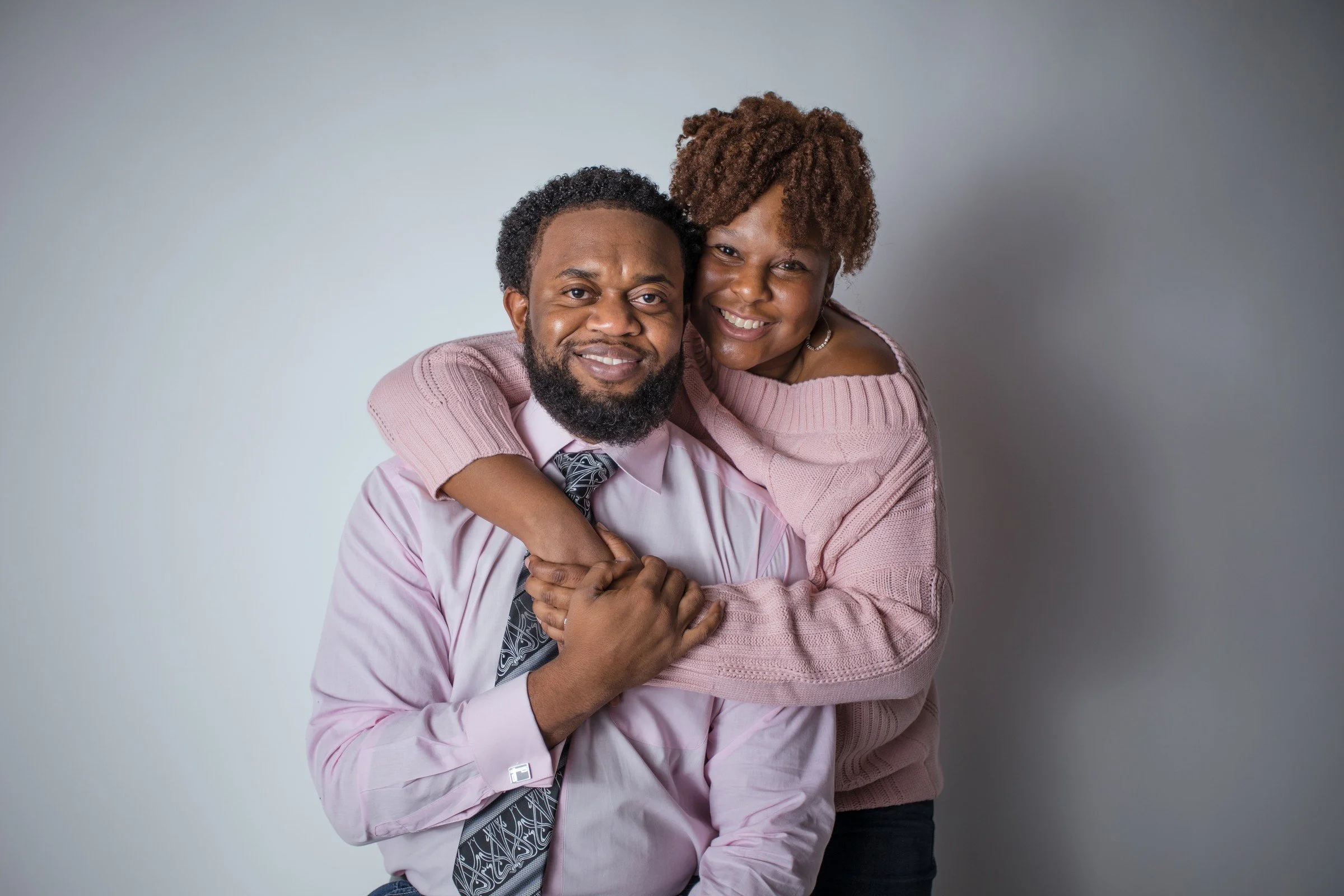 A joyful African-American couple smiling and embracing each other against a plain gray background. The woman is hugging the man's shoulders from behind, and the man is sitting, wearing a pink dress shirt and a patterned tie.