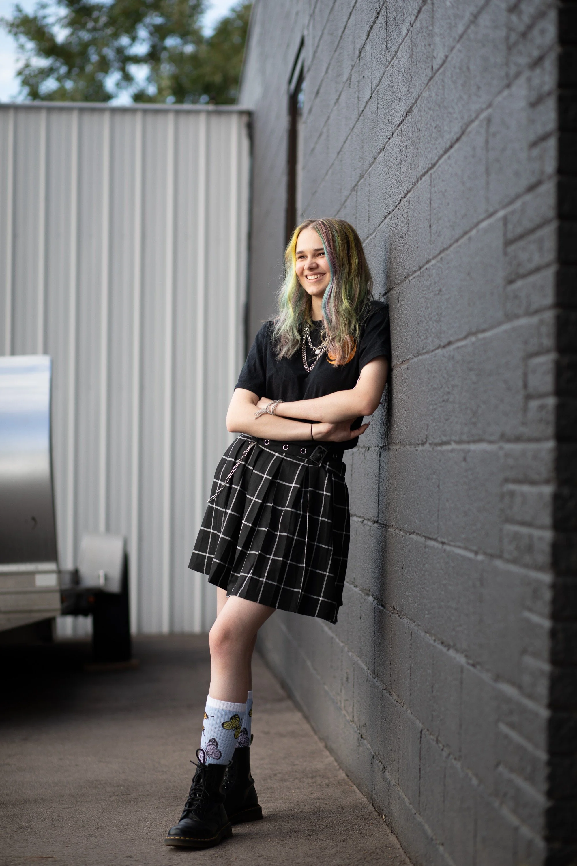 A young woman with multicolored hair, wearing a black t-shirt, black plaid skirt, white socks with butterfly patterns, and black boots, leaning against a gray brick wall and smiling.