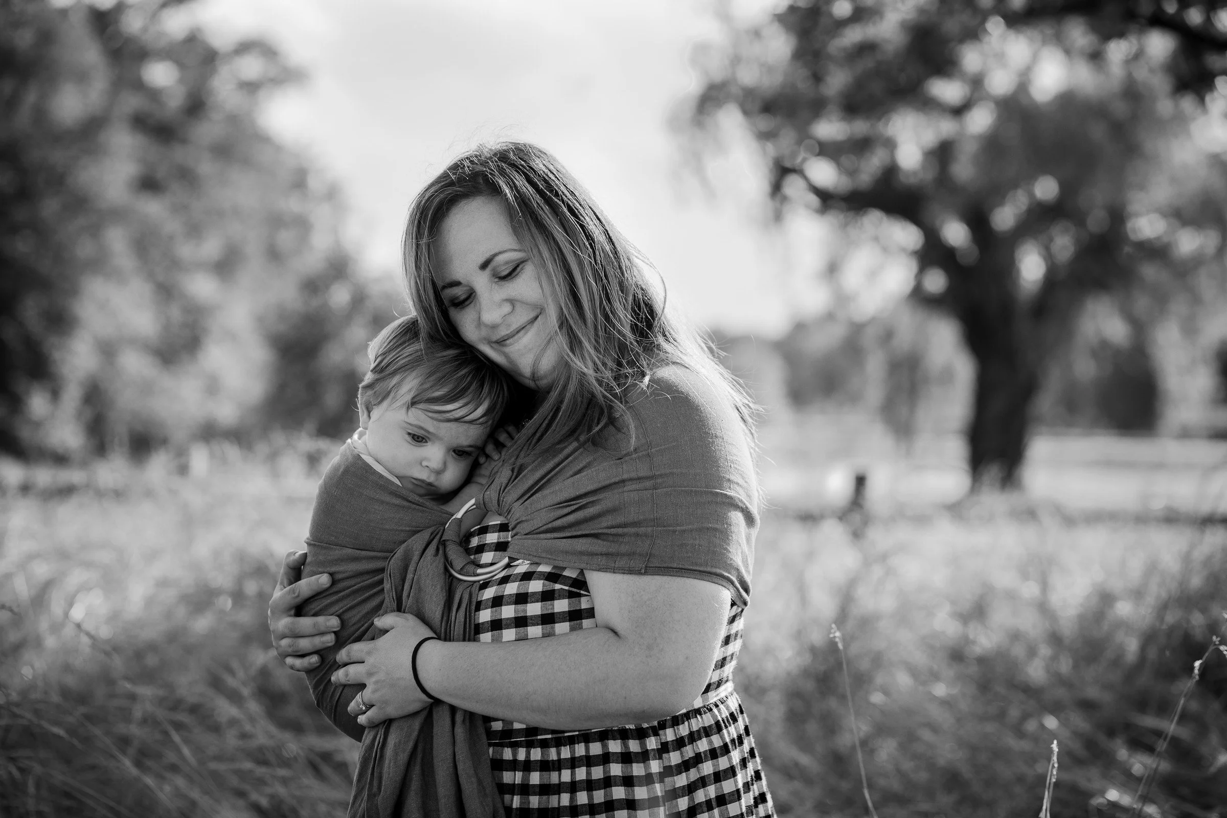A woman smiling with eyes closed, holding a young child in a baby wrap in an outdoor setting with trees in the background.