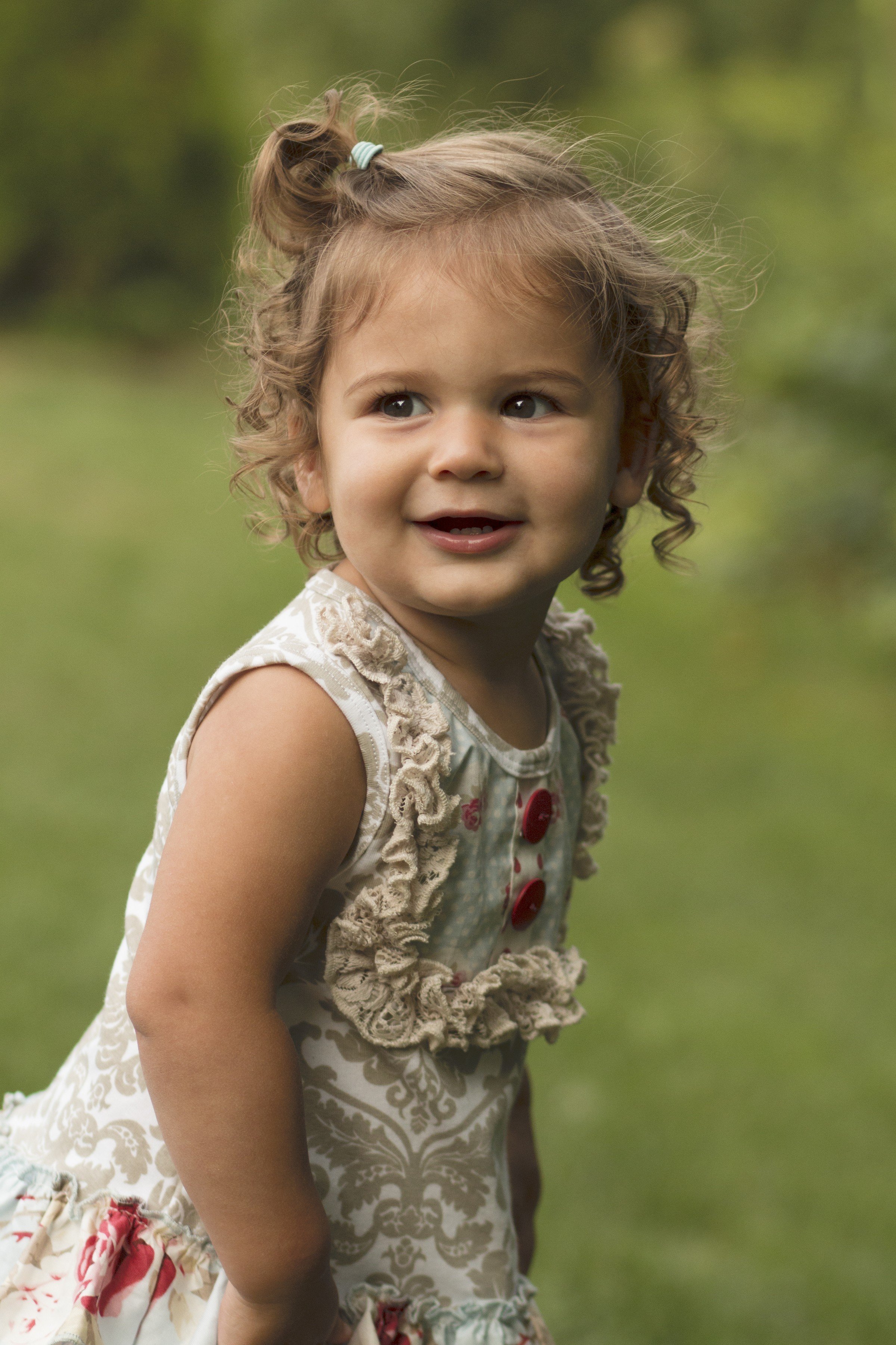 A young girl outdoors with curly hair, wearing a sleeveless dress with ruffles and red buttons, smiling and looking to the side.