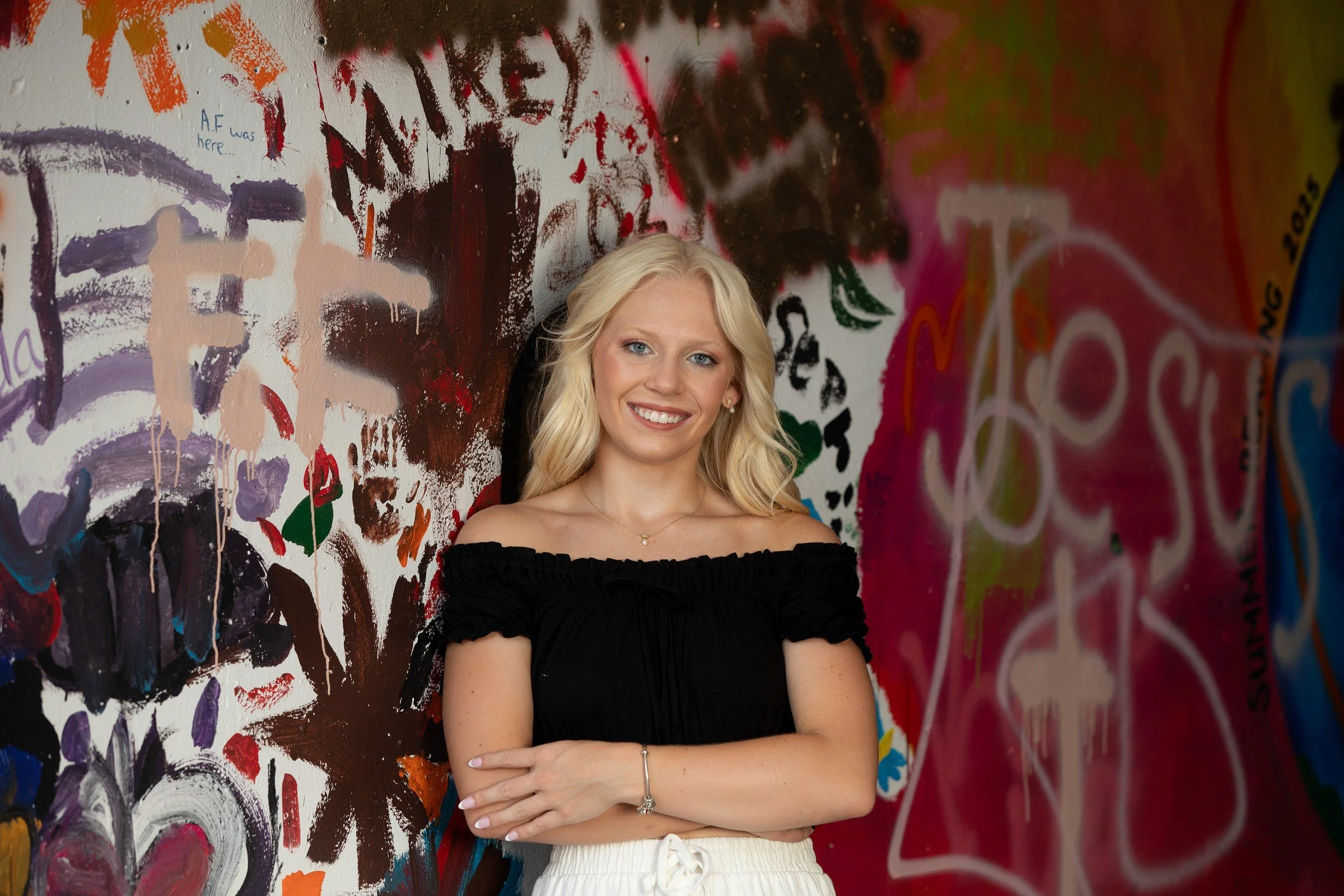 A young woman with blonde hair smiling, standing in front of a colorful, abstract graffiti wall.