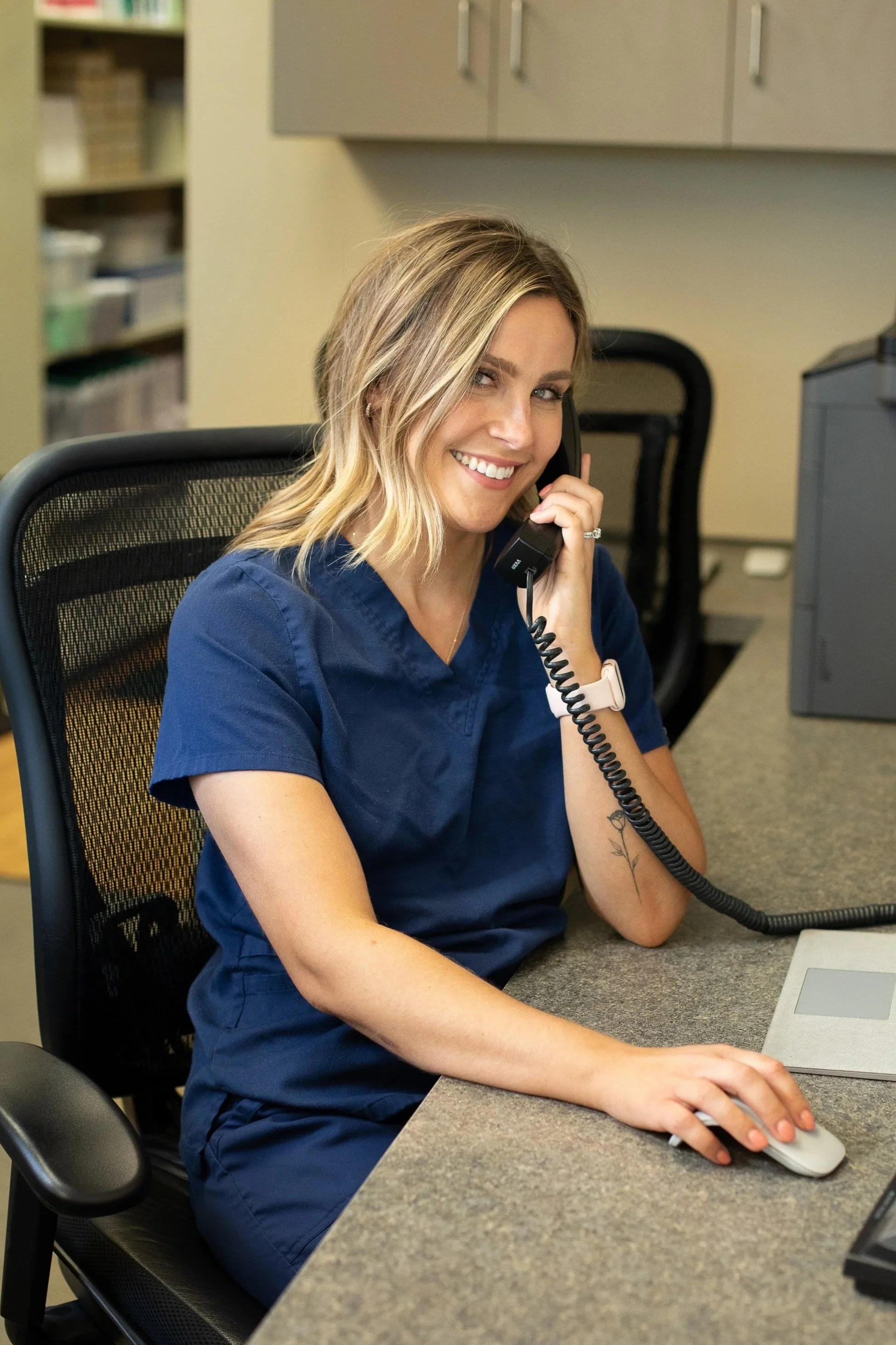 A woman in blue scrubs sitting at a desk, smiling, talking on the phone, with a computer mouse in her right hand.