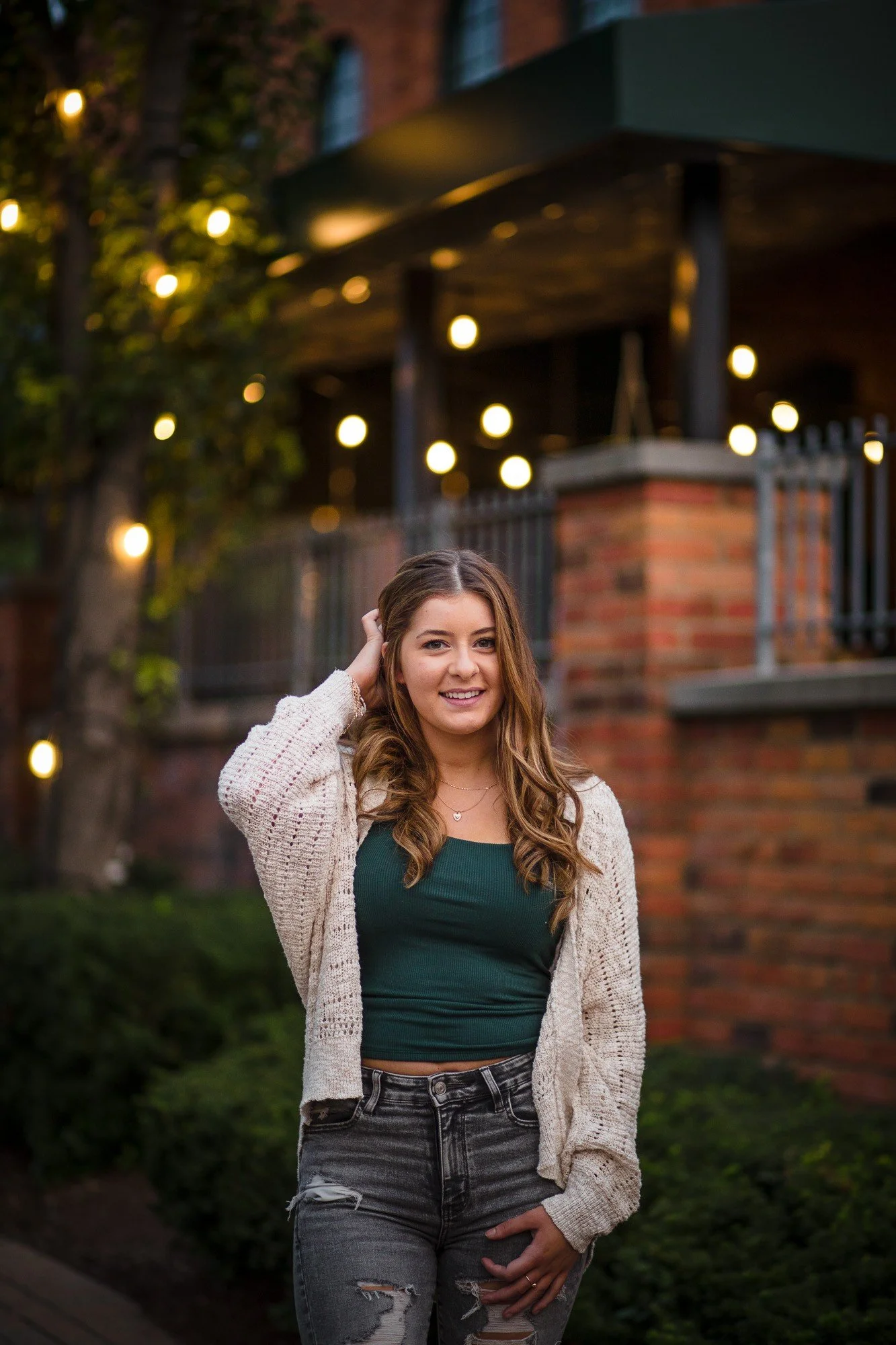 A young woman with long wavy hair smiling outdoors during twilight, wearing a green top, distressed jeans, and a beige knitted cardigan, with string lights and a brick building in the background.