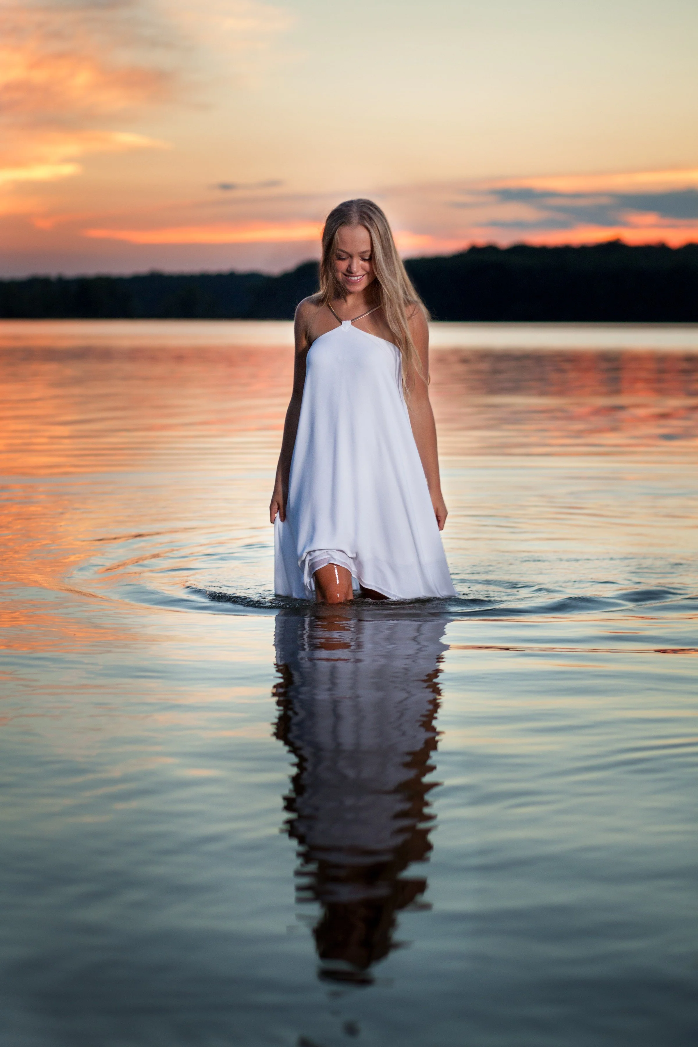 A young woman in a white dress standing in a body of water at sunset, smiling and looking down, with her reflection visible in the water.